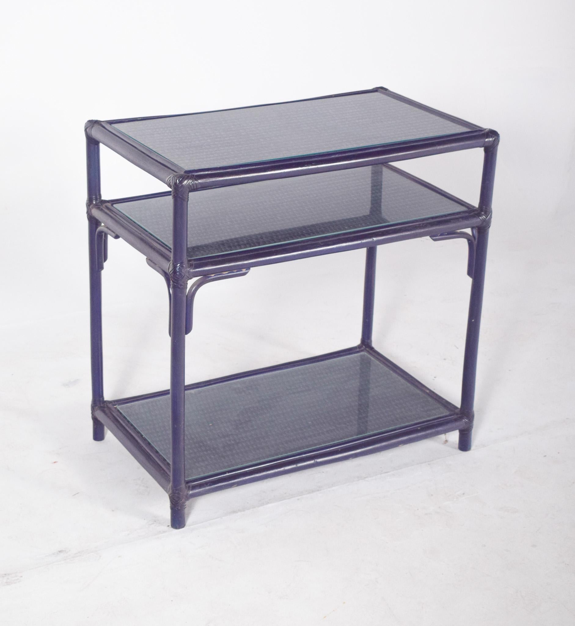 Side table in wicker, painted blue. With a light and functional structure, made up of three levels of shelves. The tubular wicker frame gives the furniture a casual, airy style, typical of tropical-inspired or balcony pieces. The dark blue paint