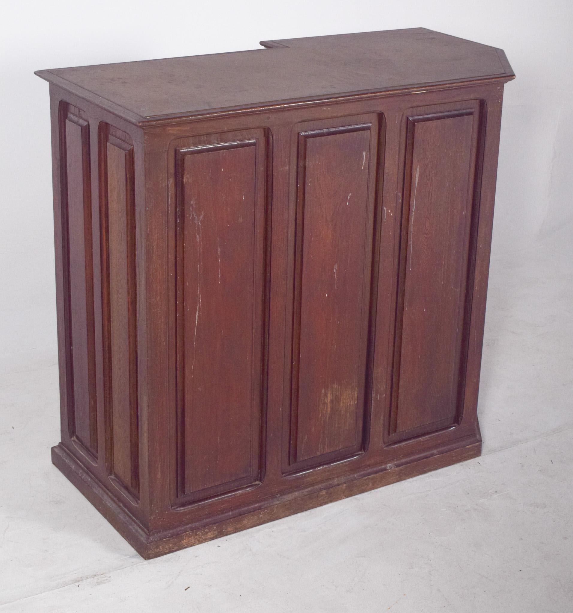 Mobile bar in legno. Con una finitura scura e robusta, la parte anteriore è chiusa, con quattro pannelli in rilievo che le conferiscono un aspetto classico e sobrio. La parte superiore presenta un'apertura sul retro che permette al corpo dell'utente