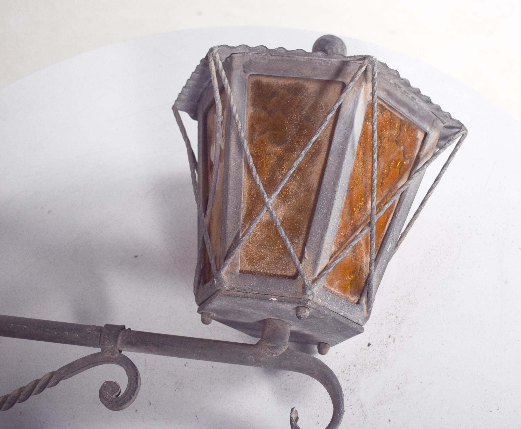 Lampione in ferro battuto e ottone. In stile rustico, è decorato con curve, foglie e spirali. La cornice ospita pannelli di vetro martellato che ammorbidiscono la luce emessa, creando un effetto diffuso e accogliente.
La lampada è sospesa da una