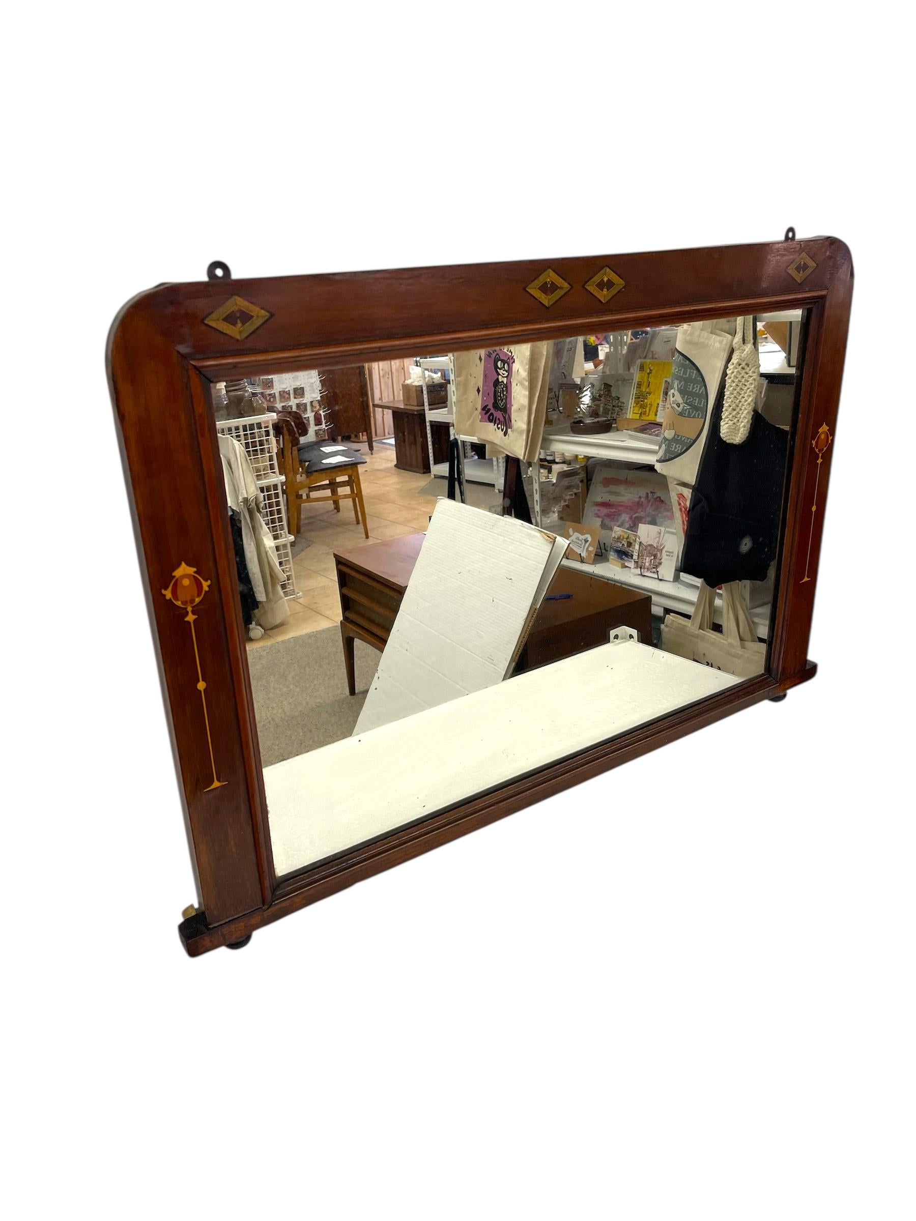 Miroir mural en acajou de style Arts and Crafts, probablement du début du 20e siècle, avec un décor peint à la main et des accents géométriques en marqueterie. Le cadre rectangulaire aux angles arrondis et adoucis est orné de détails floraux