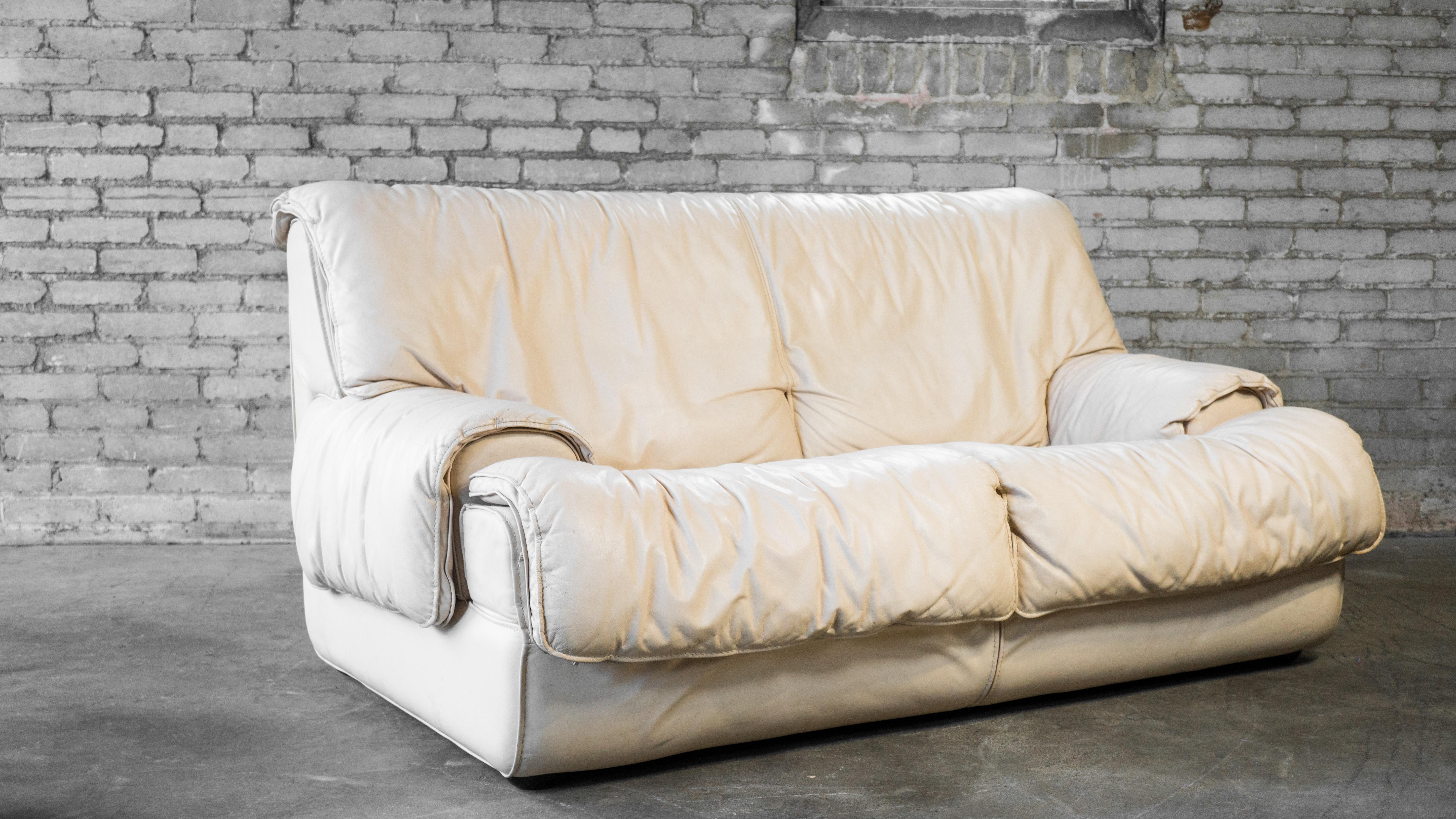 Roche Bobois cream leather loveseat, circa 1980s. Low profile design with thick, soft padding. Leather drapes over arms and backrest for relaxed vibe. Seating is very low to the ground and immersive. Good vintage condition. Manufacturer label