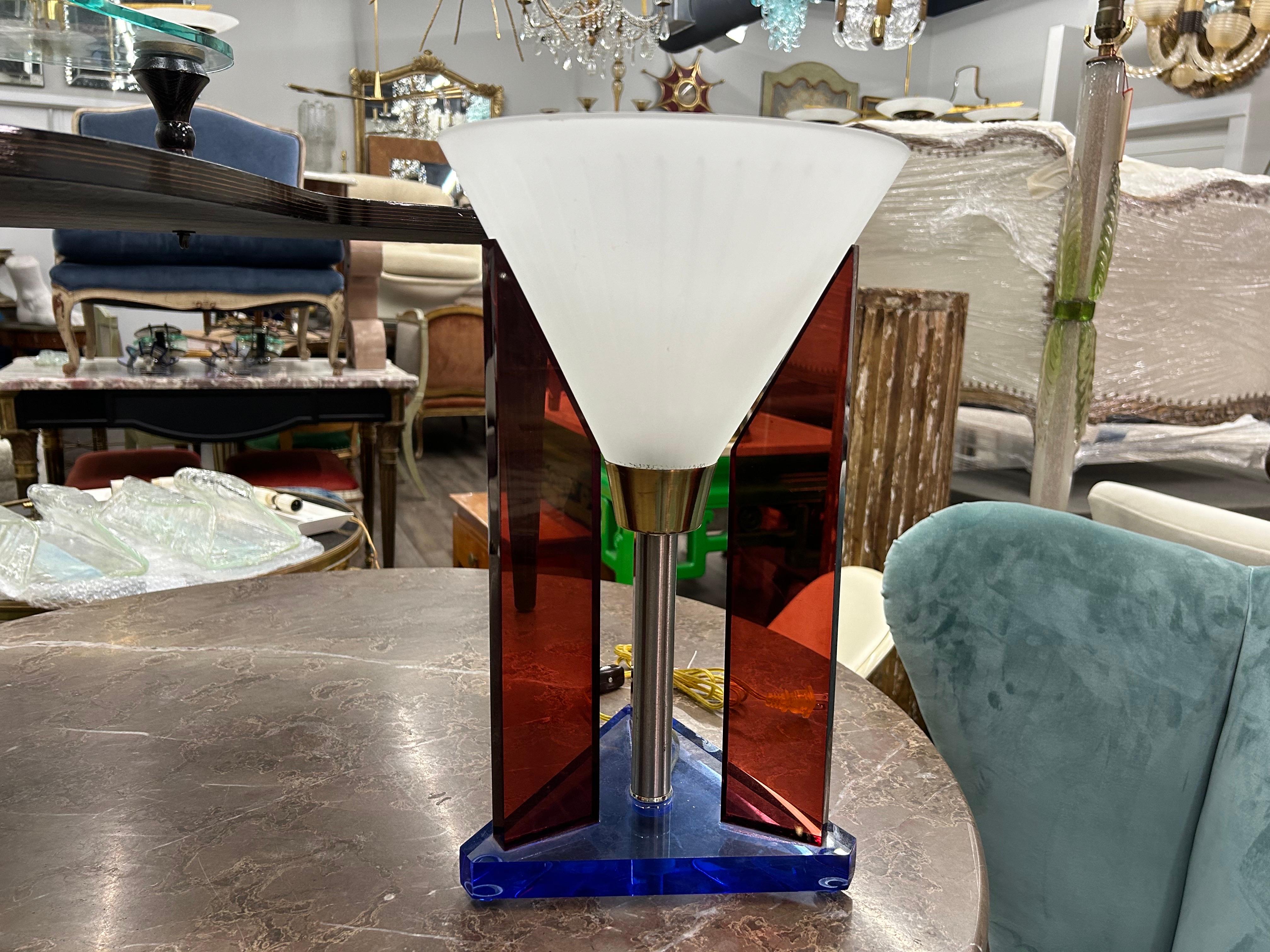 Postmoderne Lampe aus Acryl und Glas.
Diese höchst ungewöhnliche geometrische Modernist/Memphis-Lampe hat einen dreieckigen blauen Acrylsockel, der drei orange/rote Säulen mit einem zentralen kegelförmigen gestreiften Glasschirm trägt.
Unsere Lampe