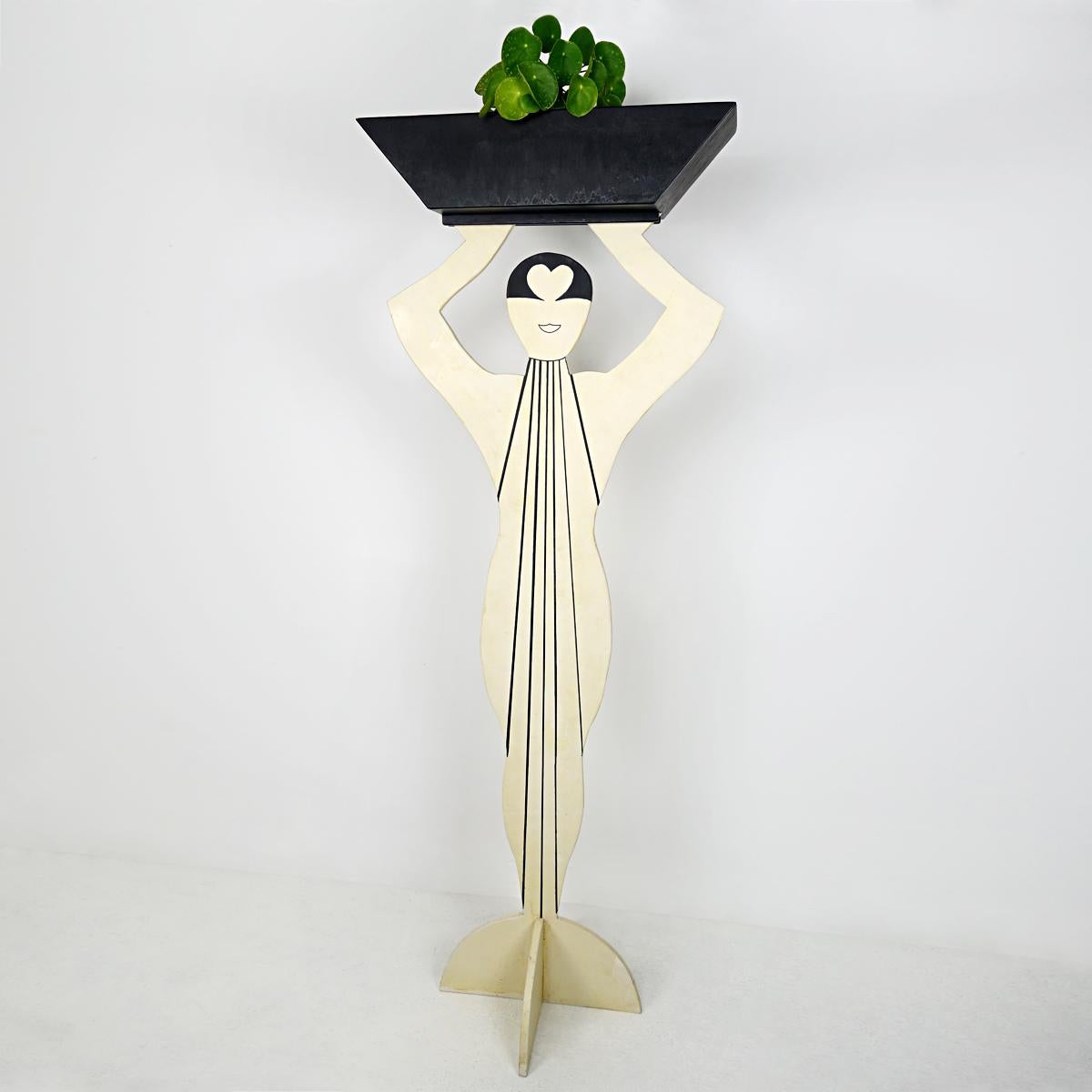 This large stylized plant stand is made of wood. It has the shape of an elegant lady dressed up in the style of the Art Deco heydays. Above her head she carries a planter that serves as the cachepot.
The object is painted on both sides so it can be