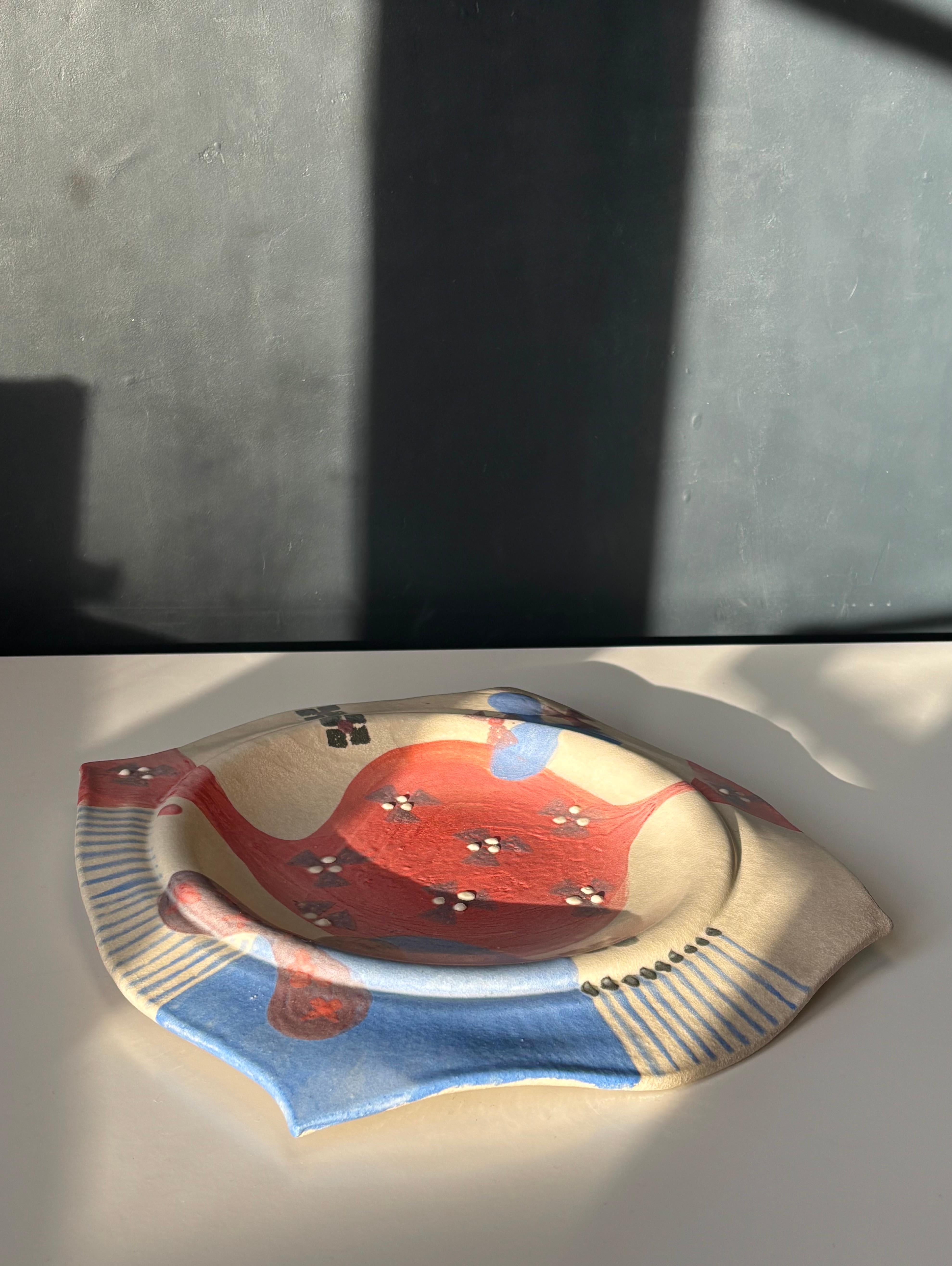 Handmade ceramic plate with organic asymmetrical shapes in muted colors. Hand-painted white, coral red and dusty blue forms and lines with small crisp white dots. Can be used as a decorative centerpiece, serving dish or as a wall plate. 
Creator