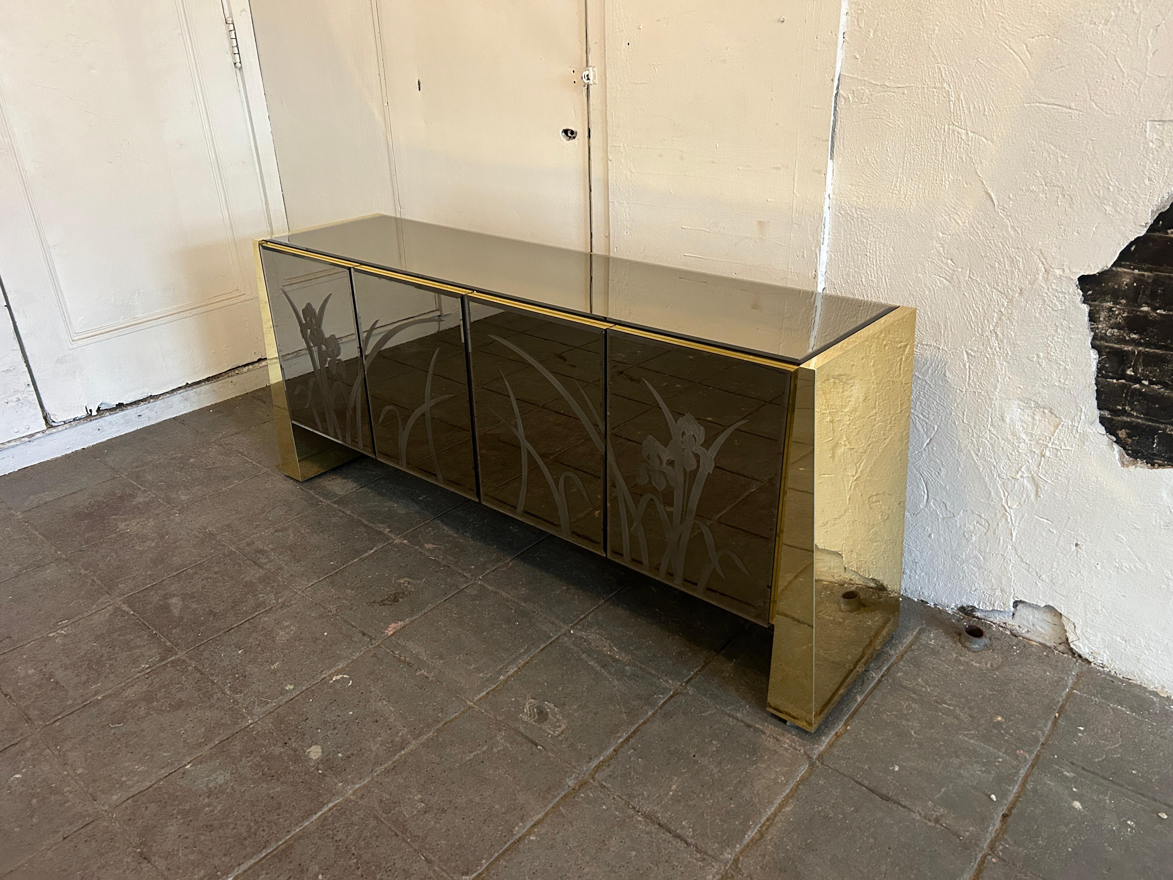 Post Modern Floral smoked mirror glass 4 door credenza with mirror brass by Ello. Great Post Modern mirror sideboard with 4 Doors adjustable shelves and (1) drawer. The front doors have floral etched pattern. The sides have Brass chrome angle legs.