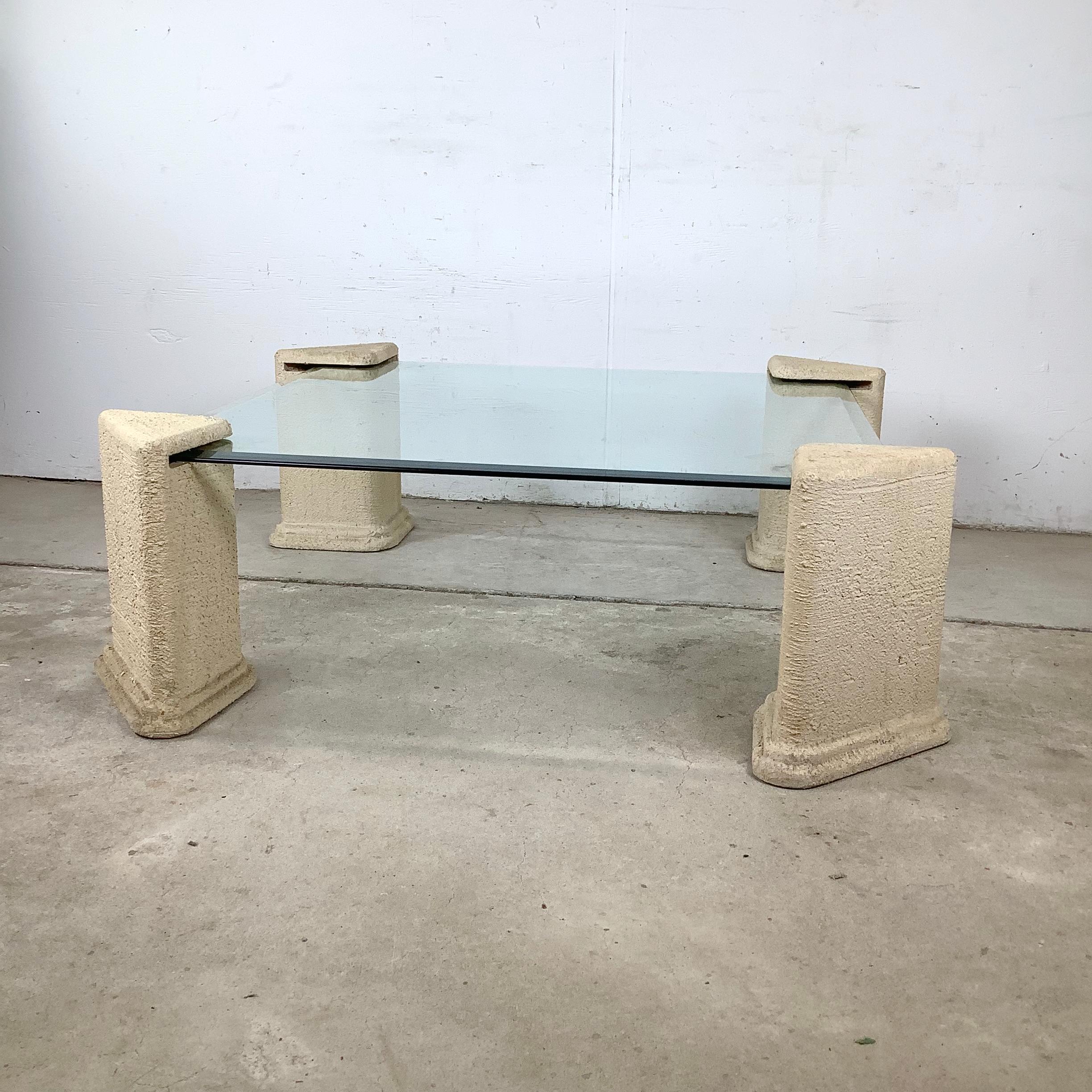 This vintage 1980s postmodern coffee table embodies the bold architectural spirit of the era. A thick square glass top floats elegantly atop four sculptural pedestal bases, each crafted with a textured stone-like finish that recalls classical