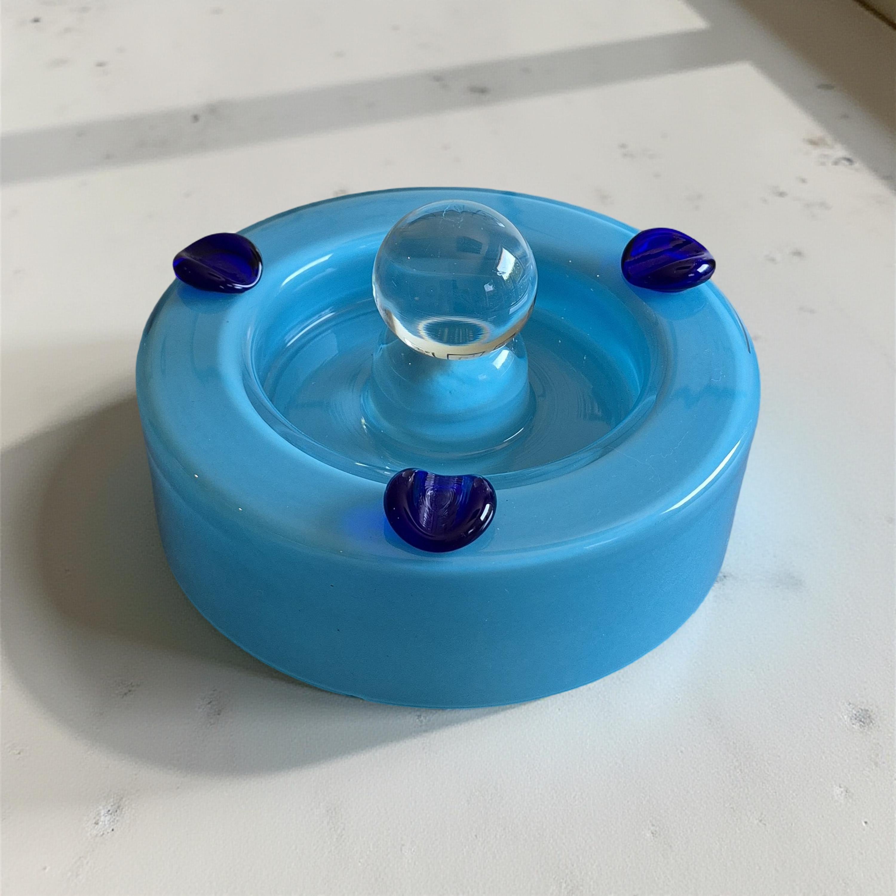 Post modern ashtray. Made from glass. The upper layer of the bowl is made from clear glass with a protruding globe on top. The inner layer of the bowl and the cigarette rests are made from blue glass.
The ashtray is in good condition. Some light