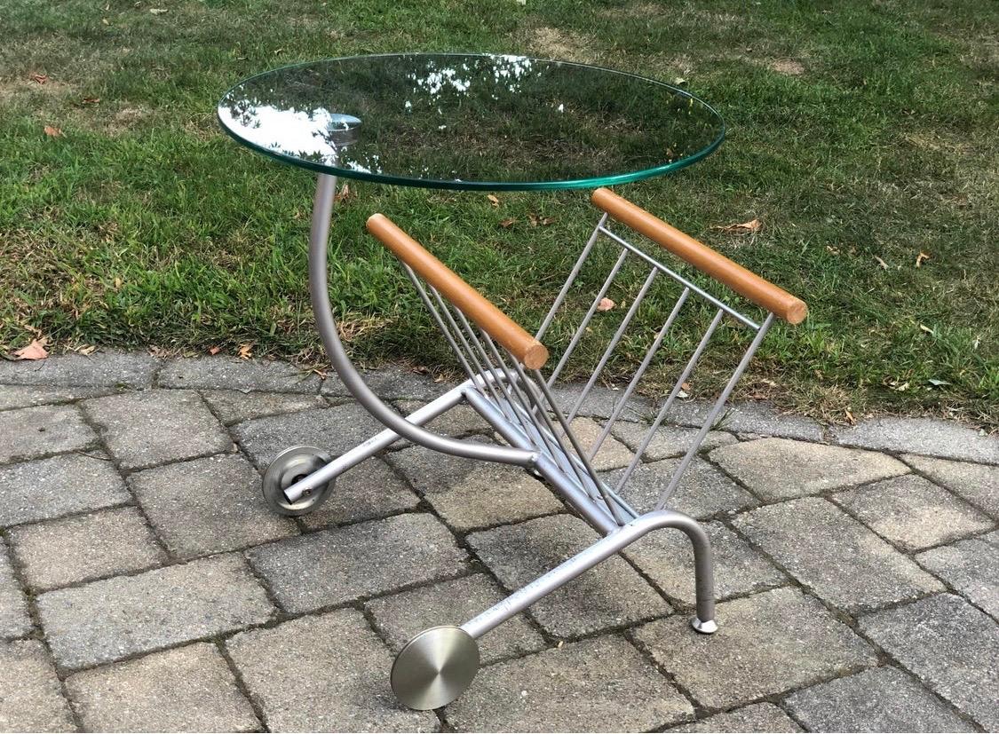 Memphis style side table with magazine holder attributed to Peter Maly for Ligne Roset. Features
glass top supported by stainless steel curved arm and frame. The magazine holder portion has wooden
brackets and functioning wheels. Age appropriate