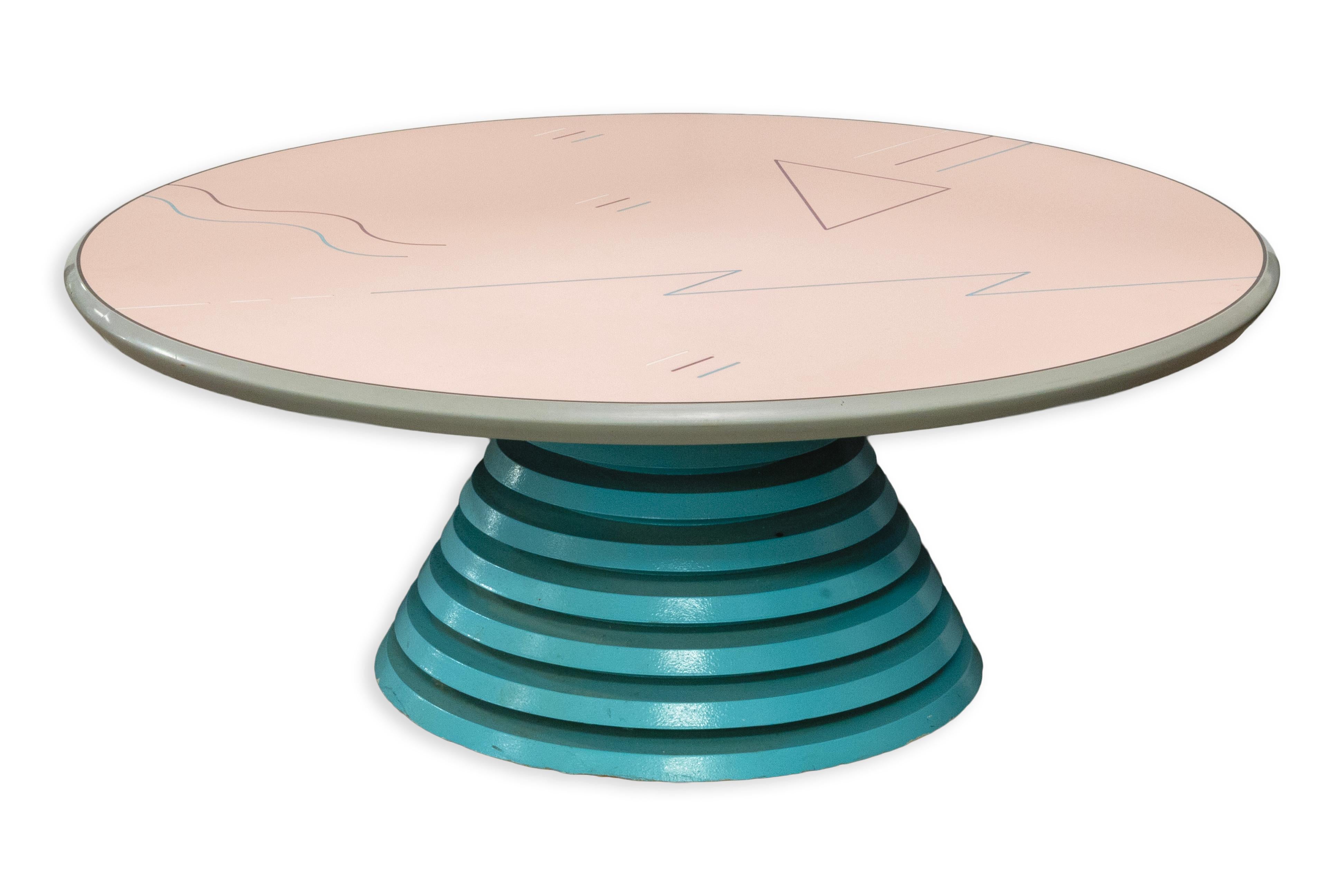 Une table basse postmoderne audacieuse dans le style de Memphis, avec une base conique empilée en laque sarcelle et un plateau circulaire en rose pastel. La surface présente des lignes abstraites ludiques et des motifs géométriques qui soulignent