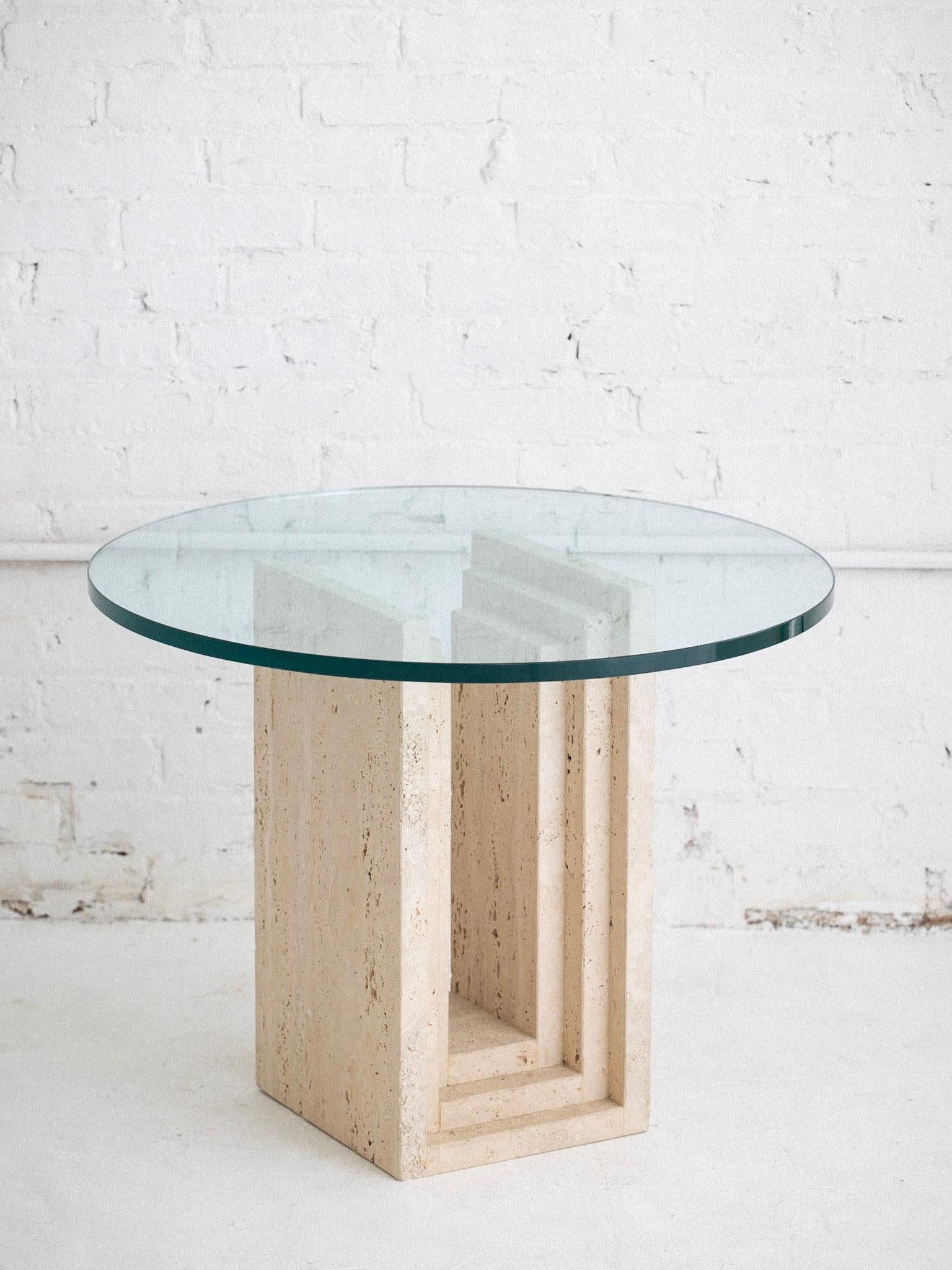 Table d'appoint post-moderne en travertin avec un plateau rond en verre. La base présente une structure étagée et superposée créant une silhouette architecturale. Le travertin brut non chargé présente une surface texturée naturelle. Solide et