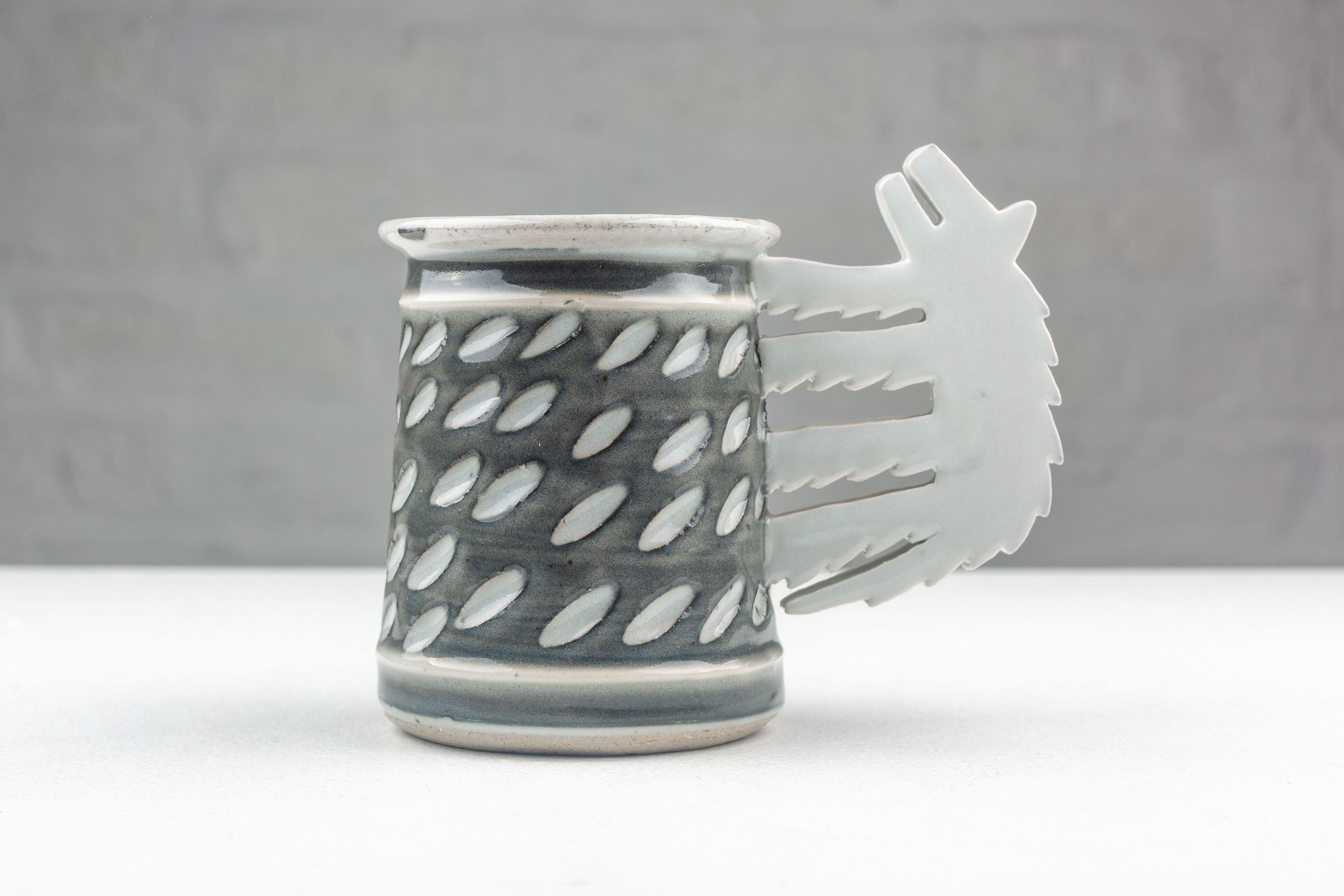 Crafted with a post-modern sensibility, this American Studio Pottery mug is the creation of Steve and Miky Cunningham, made by hand in their Iowa workshop in 1991. The piece is a study in the fusion of function and whimsy, as utilitarian form meets