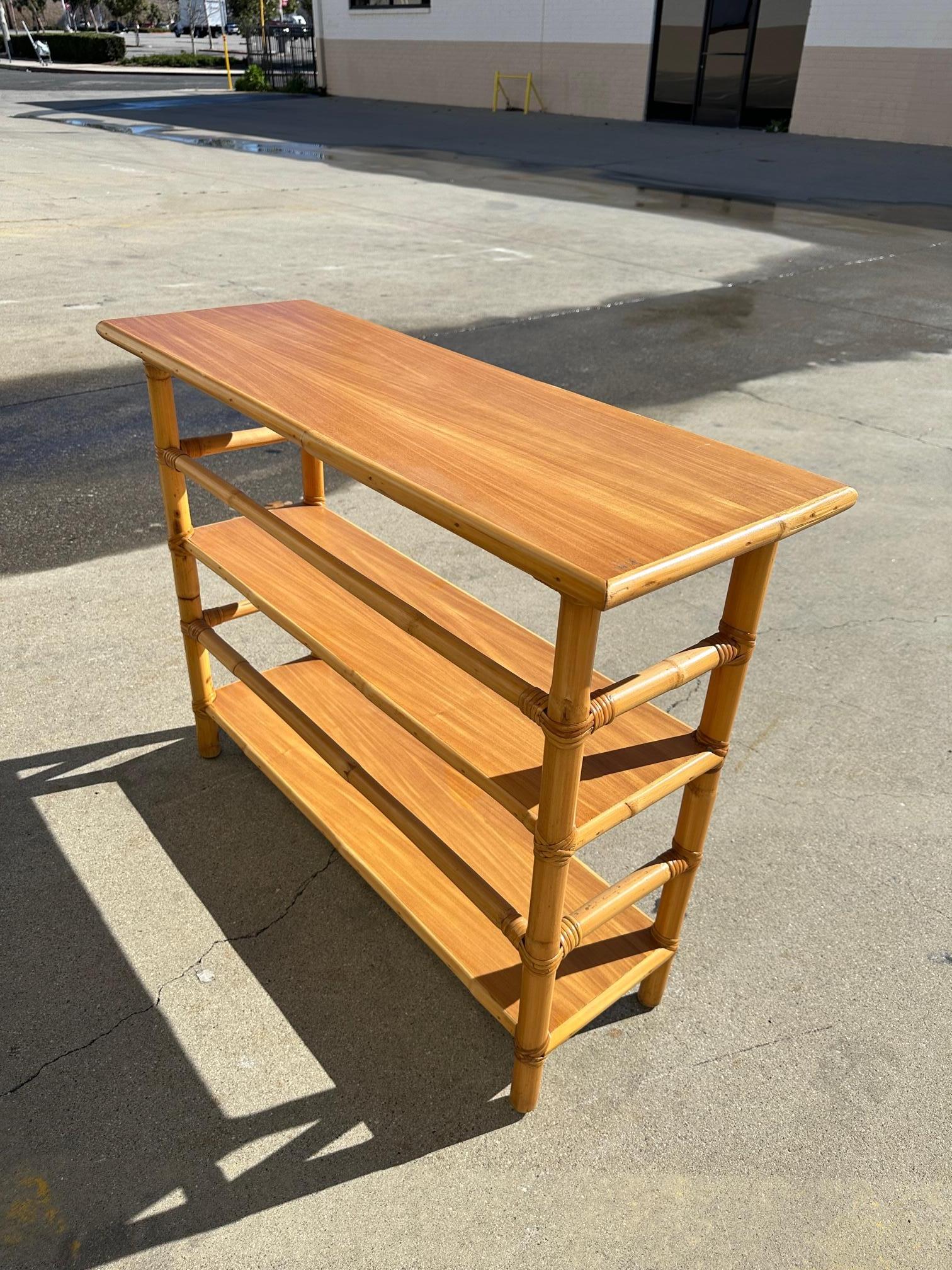 Postwar three-tier console table Bookshelf with mahogany tops and a wrapped stick rattan frame. The table has mahogany tops along each shelf which are decorated with rattan trim and each shelf has rattan pole rails for holding books in place. 

This