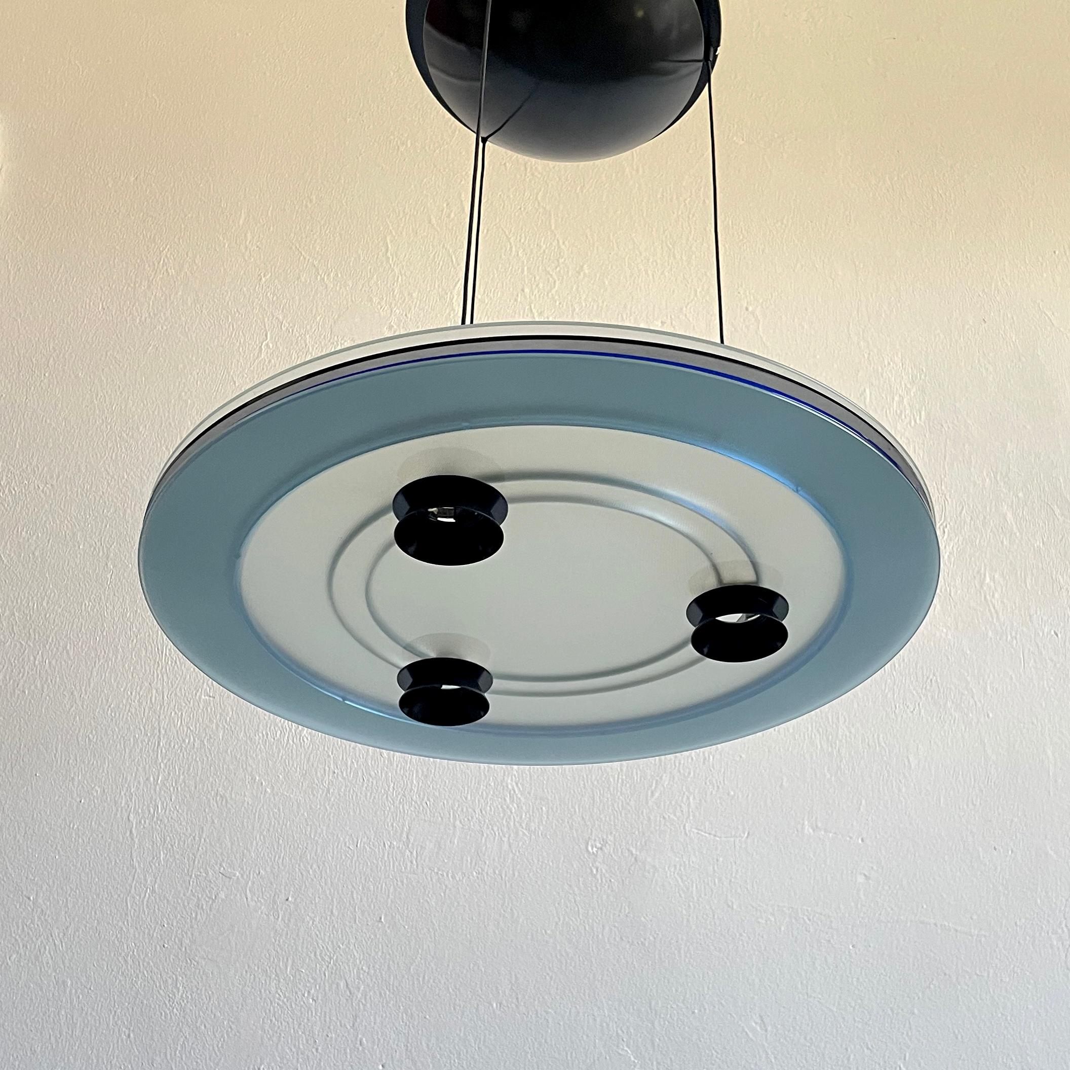 The postmodern halogen 'Aurora' chandelier light was designed by Perry King and Santiago Miranda for Arteluce in the 1980s. 
The chandelier is made of tempered glass with two acrylic transparent blue outer rings placed between. The lamp has three
