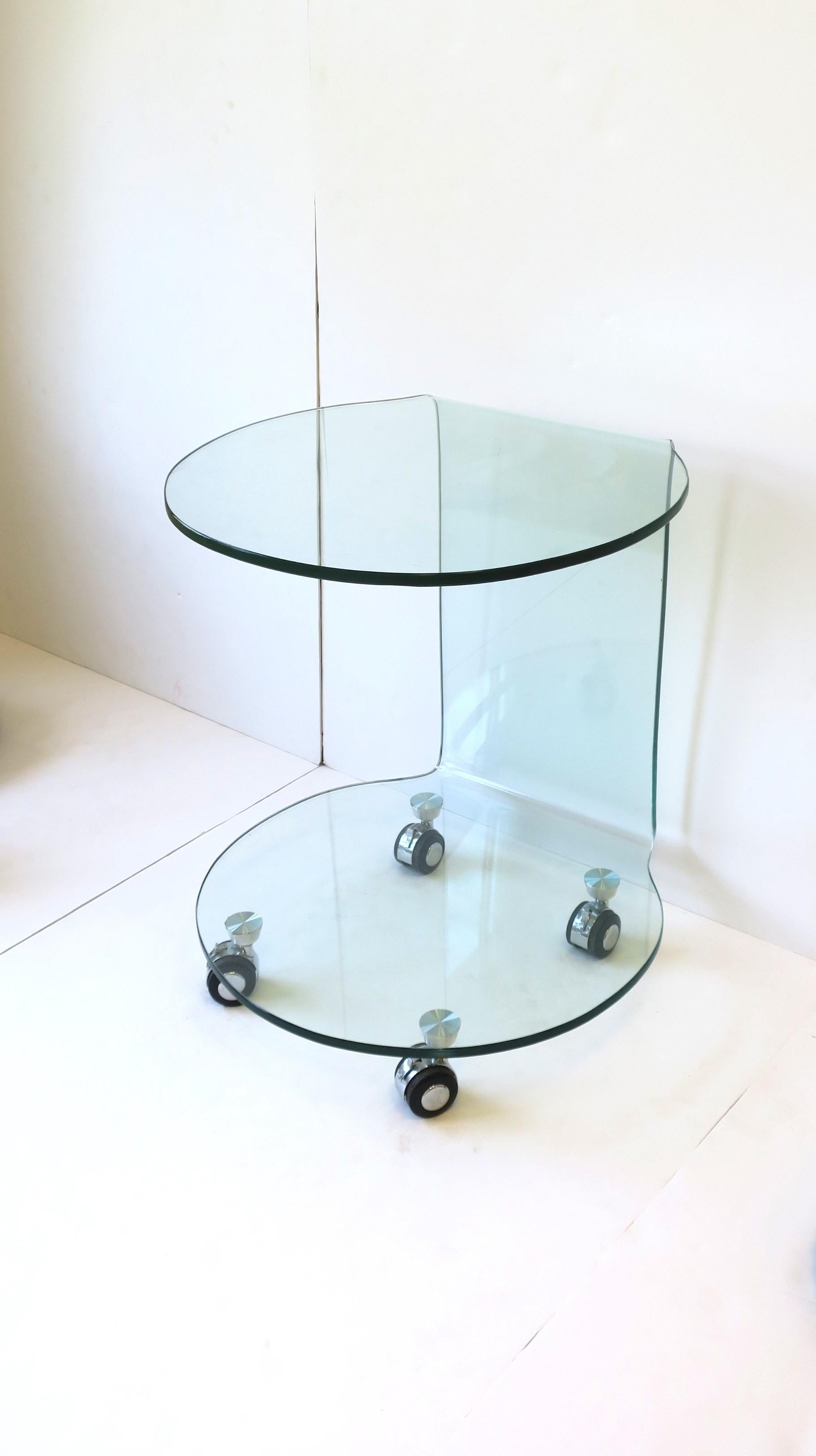 A Postmodern bent glass side or end table in the style of Italian design house Fiam, circa late 20th century. A substantial all glass, bent glass, side or drinks table, with lower shelf and caster wheels at base. Wheels do effectively roll, or