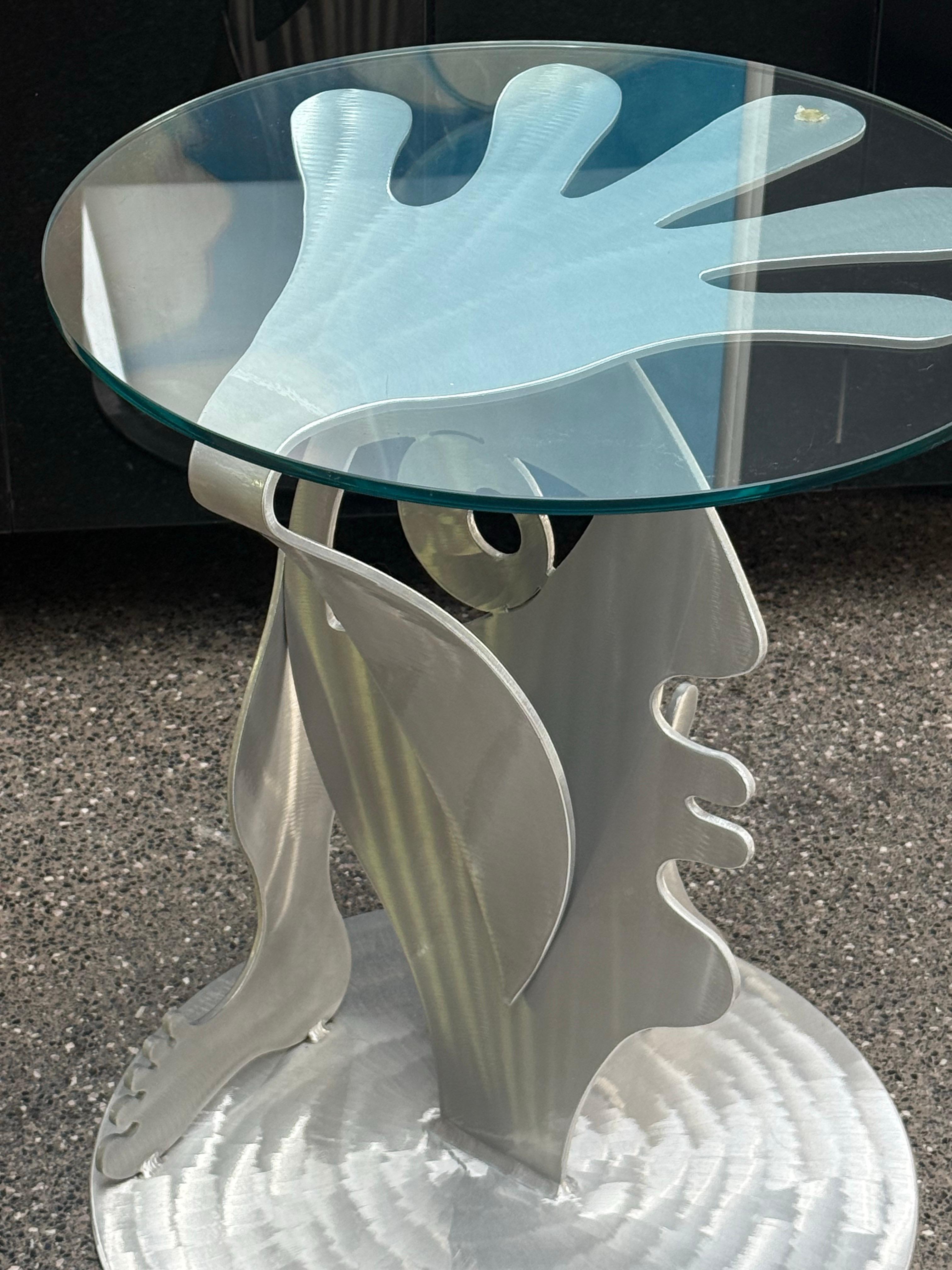 Einzelner Beistelltisch aus der Christopher Royal Collection'S für Rockledge Design Studios. Der Sockel besteht aus lasergeschnittenem Aluminium und die Tischplatte aus rundem Glas. Auf dem Sockel sind ein Gesicht, ein Fuß und Hände abgebildet.