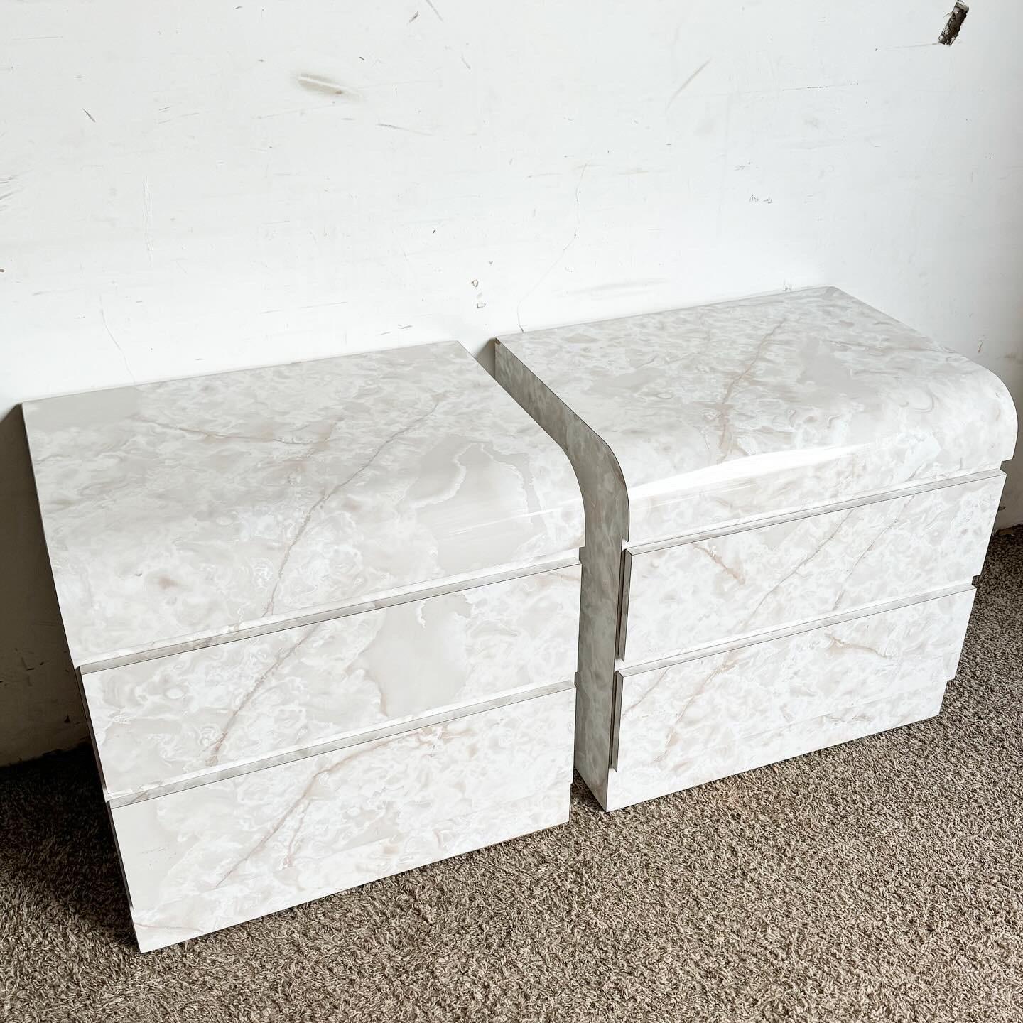 Introduce modern luxury to your bedroom with this pair of Postmodern Faux Marble Laminate Waterfall Nightstands. These nightstands feature a chic faux marble surface and a unique waterfall silhouette, combining style with practicality. With spacious