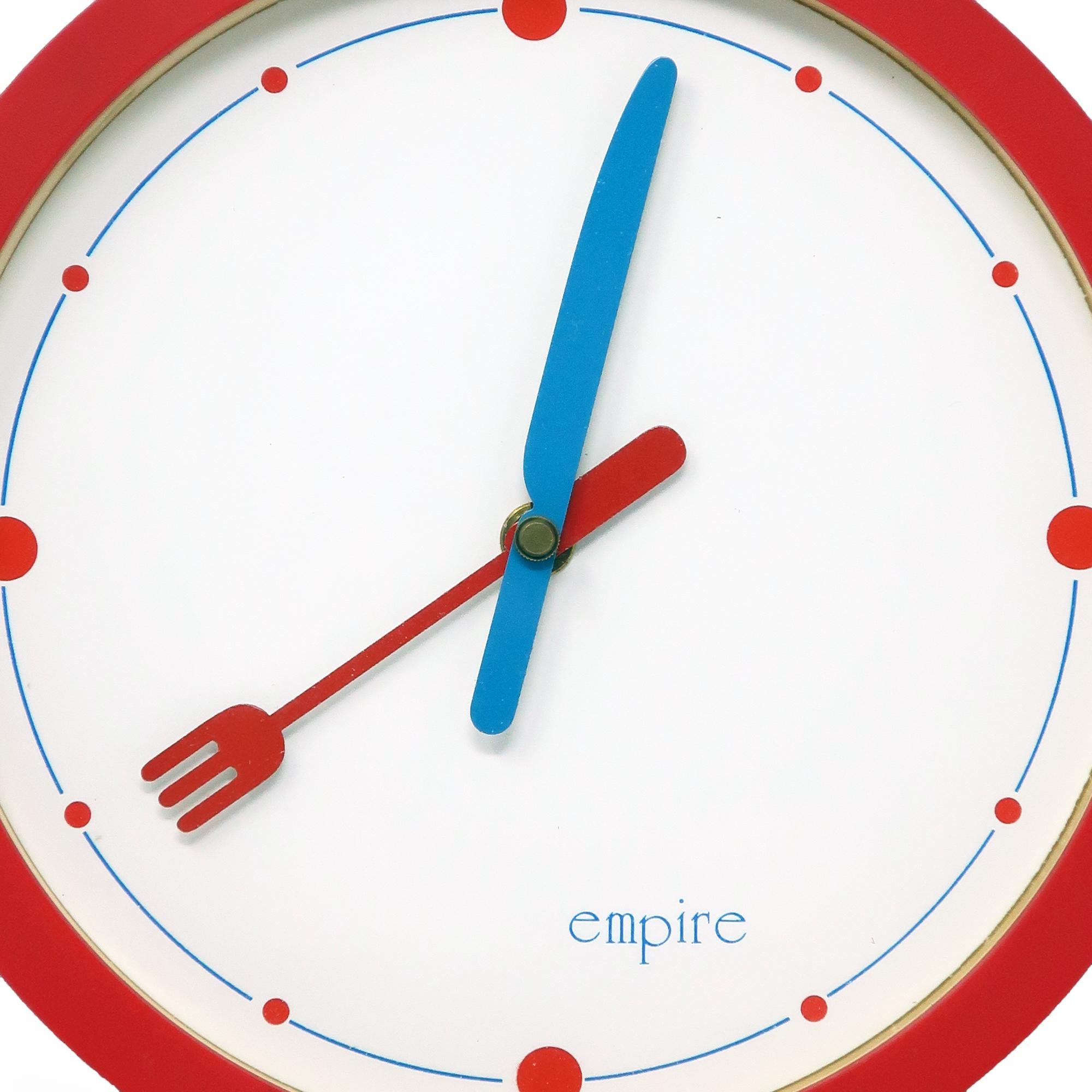 A round 1980s postmodern kitchen wall clock by Empire Arts with a red body, white face, blue and red accents, and knife and fork for minute and hour hands. A fun and whimsical wall clock that takes a single AA and works great.

In good vintage