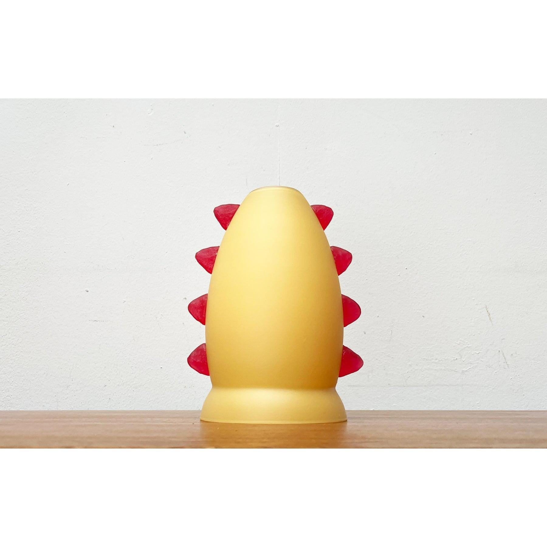 Postmodern table lamp made of glass.

A playful design. The shape and color make this small lamp appear like a living character. The glass lamp body is overlaid in yellow-orange. The glass tapers towards the top and forms a base at the bottom. Red