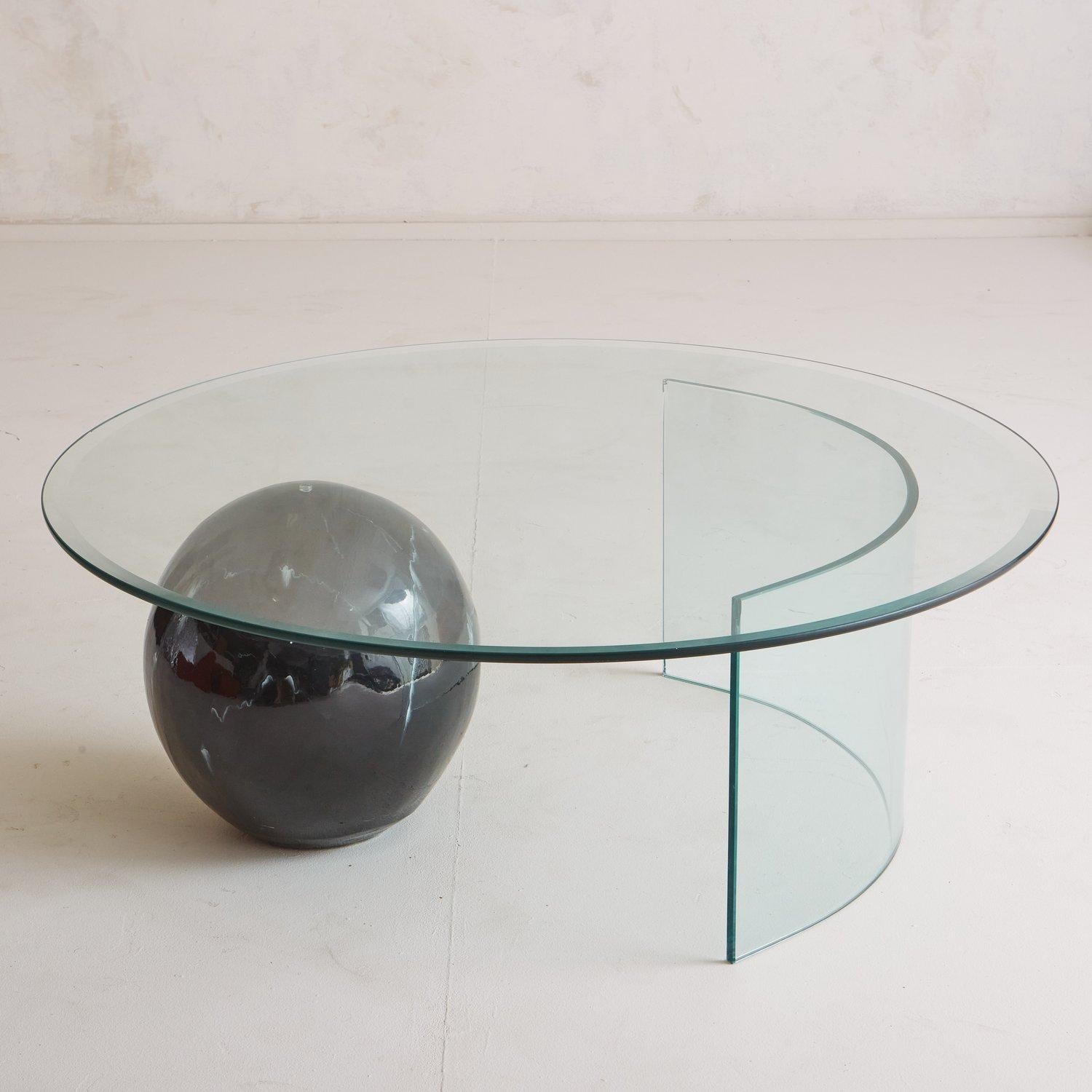 A Postmodern glass coffee table featuring a round .375” thick beveled glass tabletop resting on a black ceramic ball with a marble effect finish and a curved glass base. USA, 1980s.

 