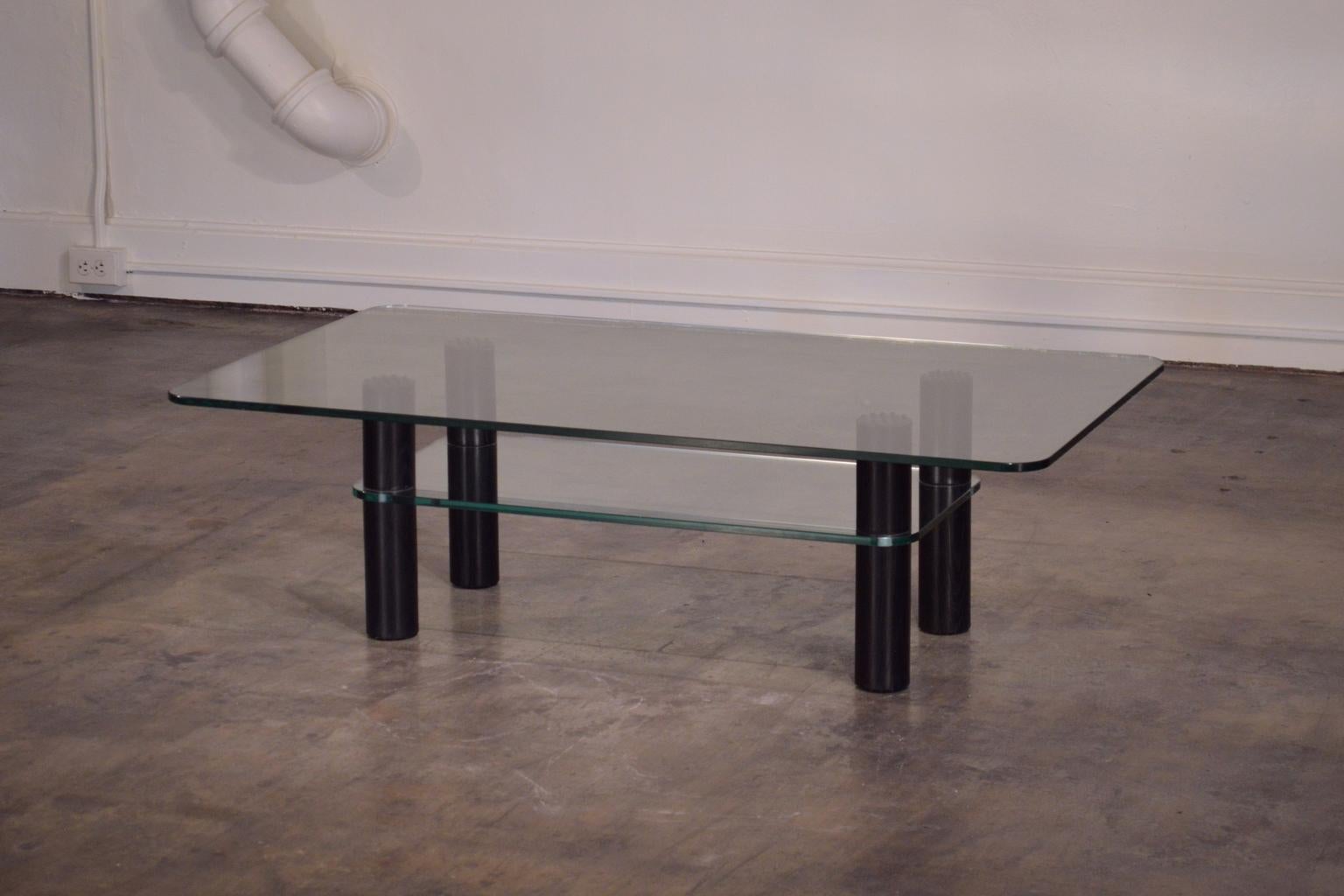 Attributed to Alessi, this sleek Postmodern coffee table features black stained oak legs, thick floating glass top, and adjustable glass shelf. The glass is edged with a very Fine bezel cut and has softened round corners. The legs unscrew and are