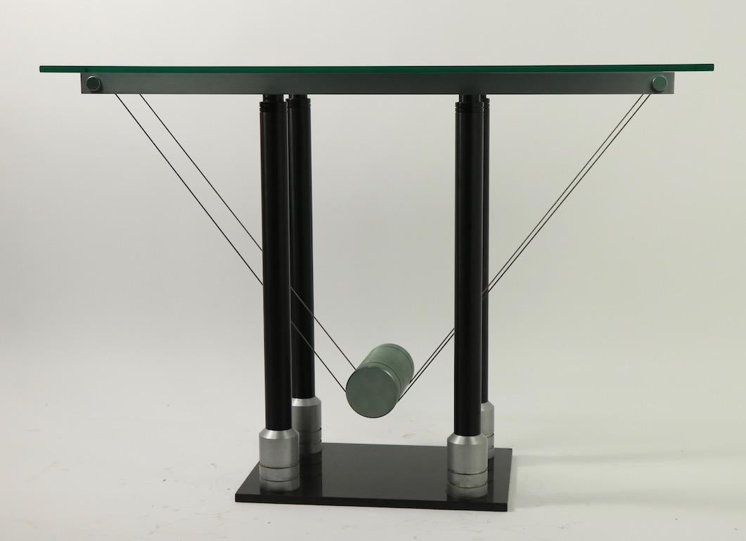 Postmodern glass top console table with suspended metal weight base. Thick glass top, four metal columns, counterweight design element and polished stone base, well constructed with top quality materials. The table is in very good condition, clean