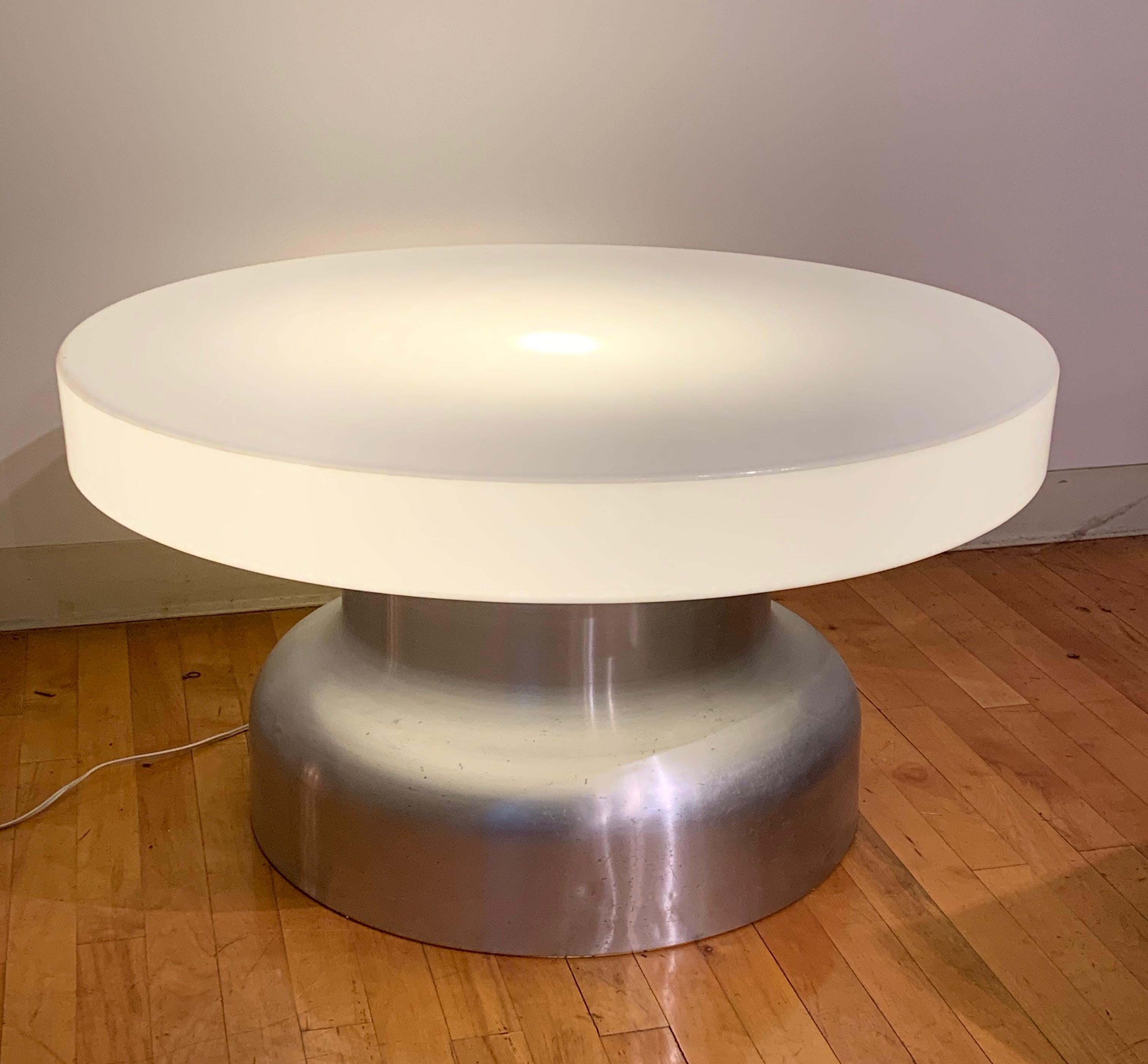 This coffee table from the 1970s has an acrylic top with a brushed aluminum cylindrical base. There is a single lightbulb located in the top. 