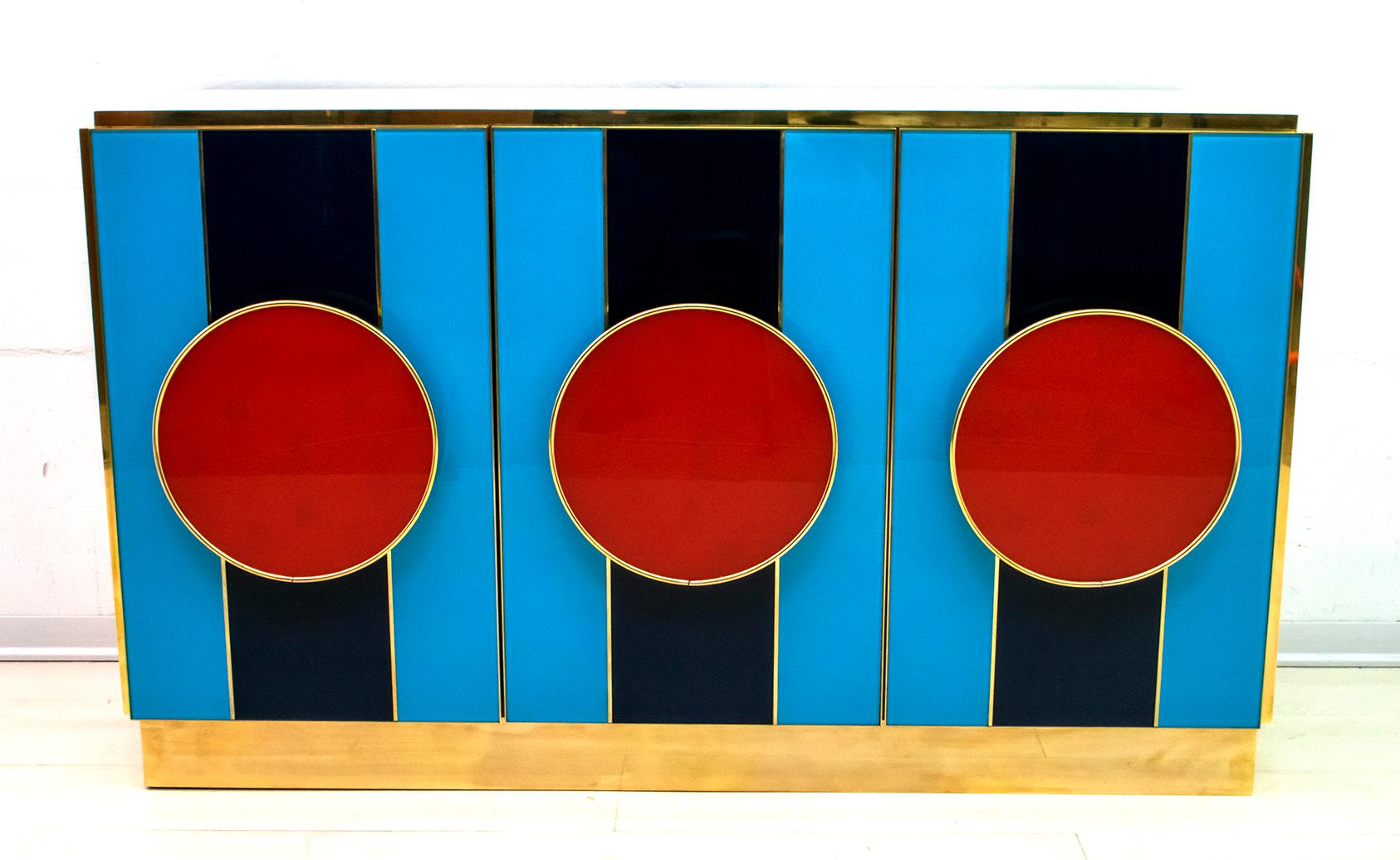 An interesting colorful Postmodern piece with a Pop Art touch, sideboard / bar cabinet, very fun Italian, design with 3 front doors in blue and light blue glass, like handles two circles in red glass, which highlight the brass finishes. The rest of