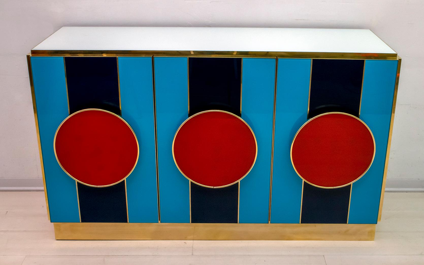 An interesting colorful Postmodern piece with a Pop Art touch, sideboard / bar cabinet, very fun Italian, design with 3 front doors in blue and light blue glass, like handles two circles in red glass, which highlight the brass finishes. The rest of