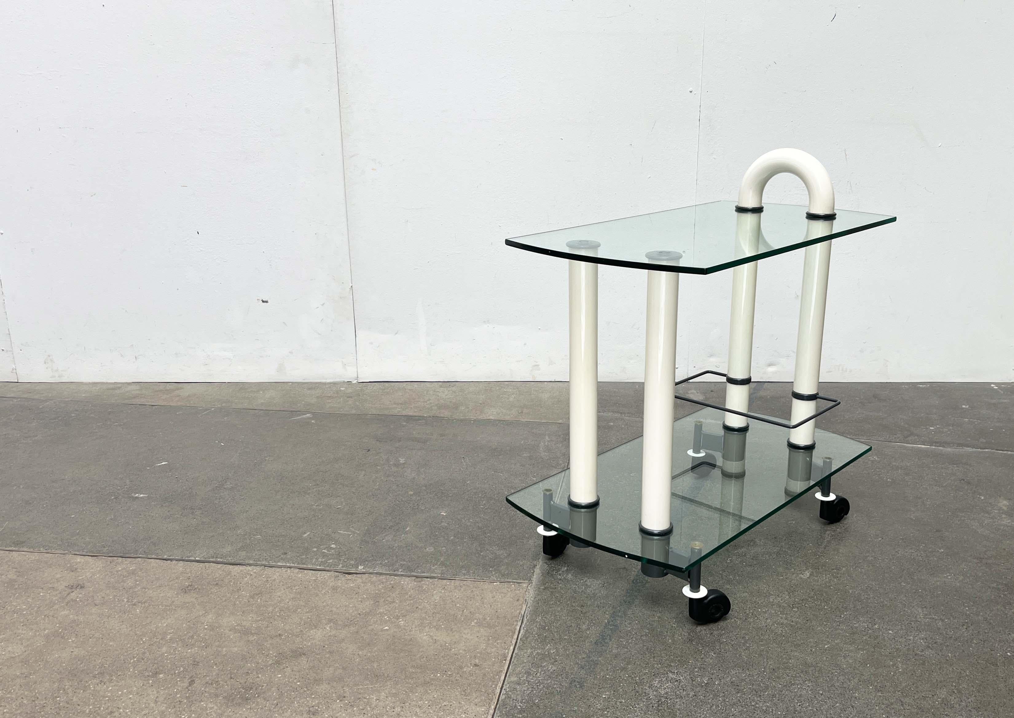 Postmodern bar cart from the 1980s by the Italian manufacturer Metalnova.

The design impresses with its clear geometry and themes of transparency and reflection. The two glass shelves rest on four white metal cylinders. The two rear supports appear