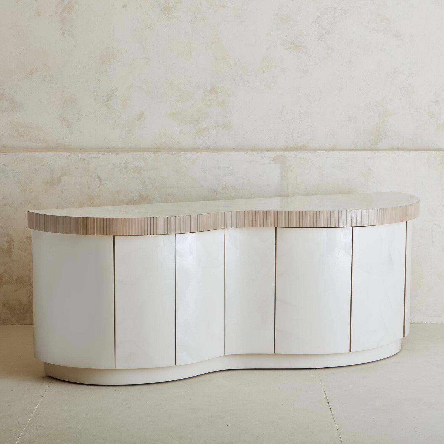 A glossy ivory laminate credenza featuring a dramatic curved front, a recessed plinth base and a taupe fluted glass tile trim. This credenza has four vertical doors that open outwards with push latch hardware, revealing four adjustable shelves and