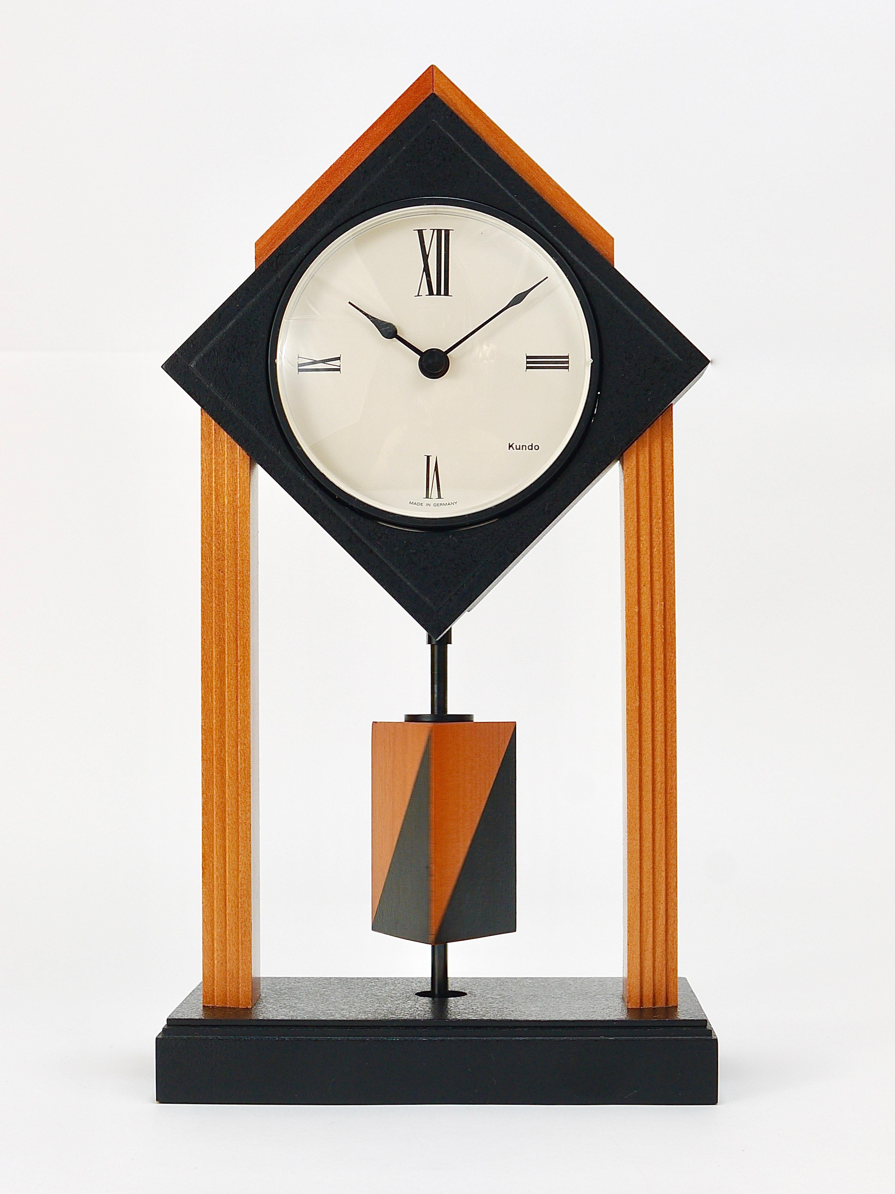 A wonderful post modern Memphis style desk or table pillar clock with a rotating triangular pendulum. Executed by Kundo Germany in the 1980s. 
Made of natural and black lacquered wood with a beautiful clocks face and nice black hands. Powered by a