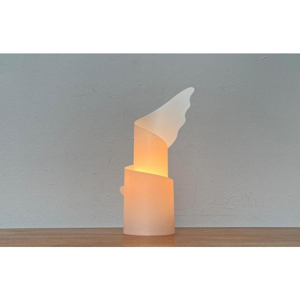 Lámpara de mesa postmoderna de Slamp según un diseño de Riccardo Raco de principios de los 90.

El cuerpo de la luminaria sirve de difusor y está formado por el plástico translúcido opalflex. El proyecto se basa en la transformación del plástico
