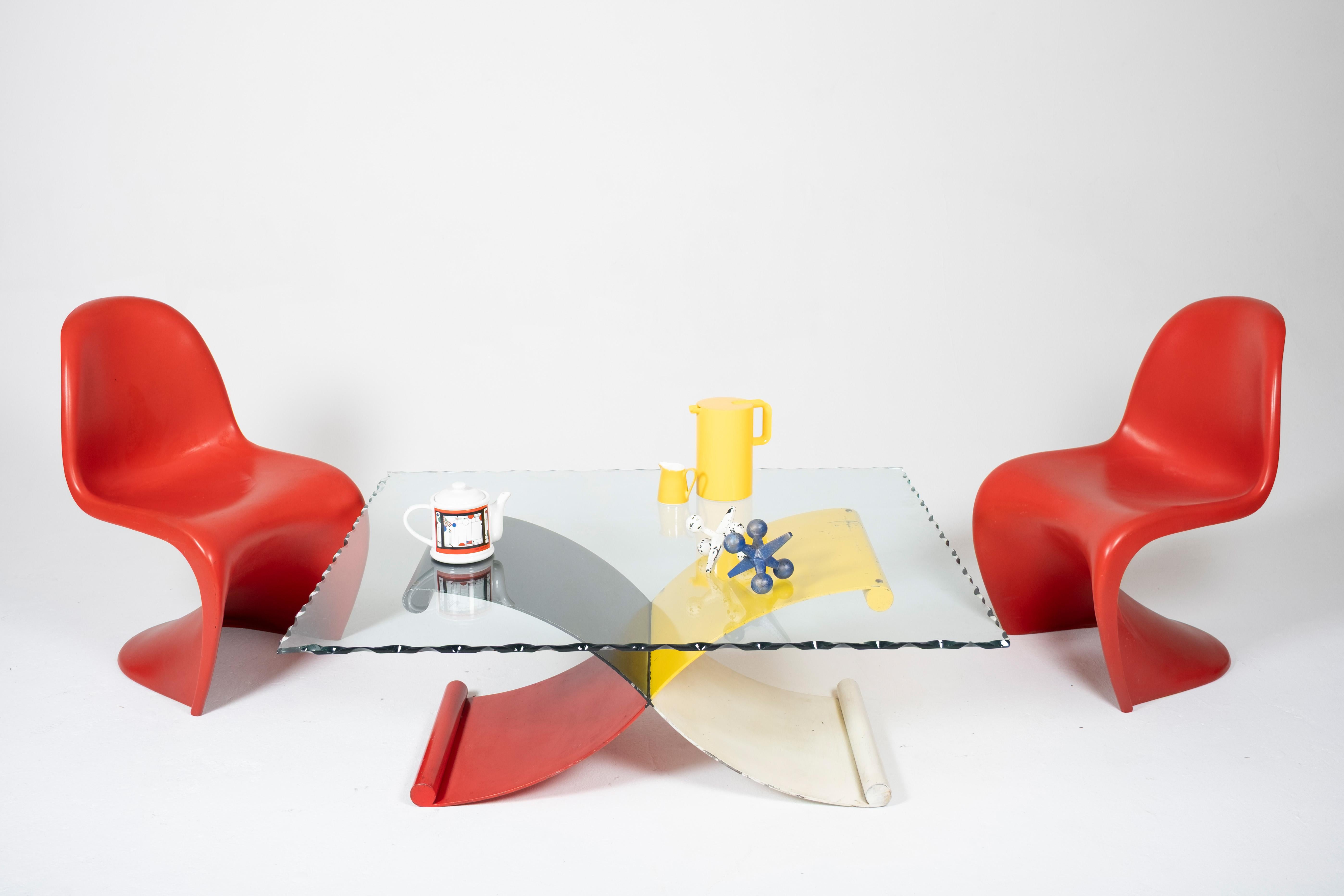 A dynamic postmodern coffee table base constructed from four intersecting curved iron forms, each painted in a bold primary tone—red, black, yellow, and white. The sculptural X-shaped design is visually engaging from every angle, offering both