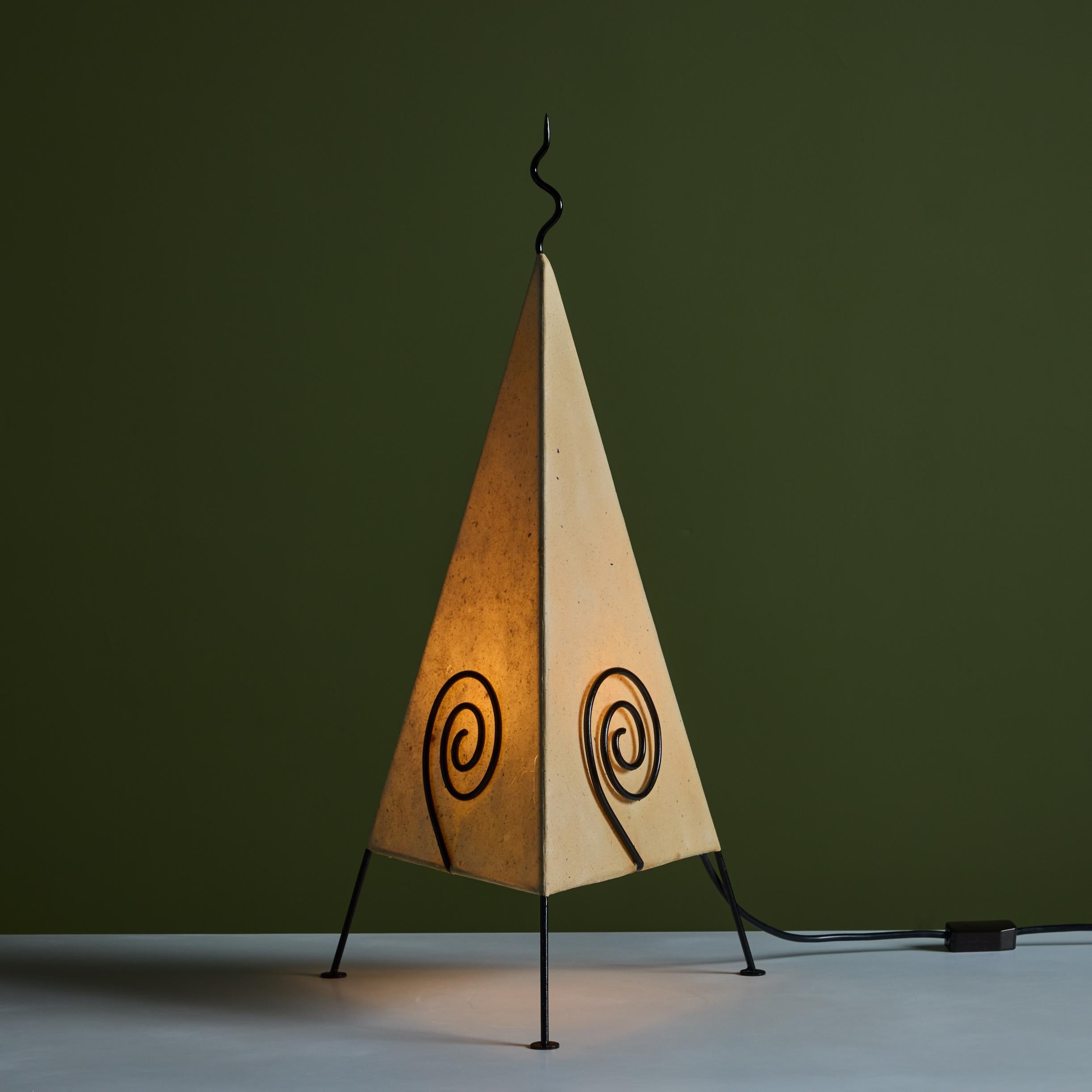 Tripod postmodern paper shade table lamp with black enameled frame. The table lamp showcases a triangular paper shade with enameled metal swirl detail on each side of the shade with a metal squiggle at the top. On/off switch is located on the