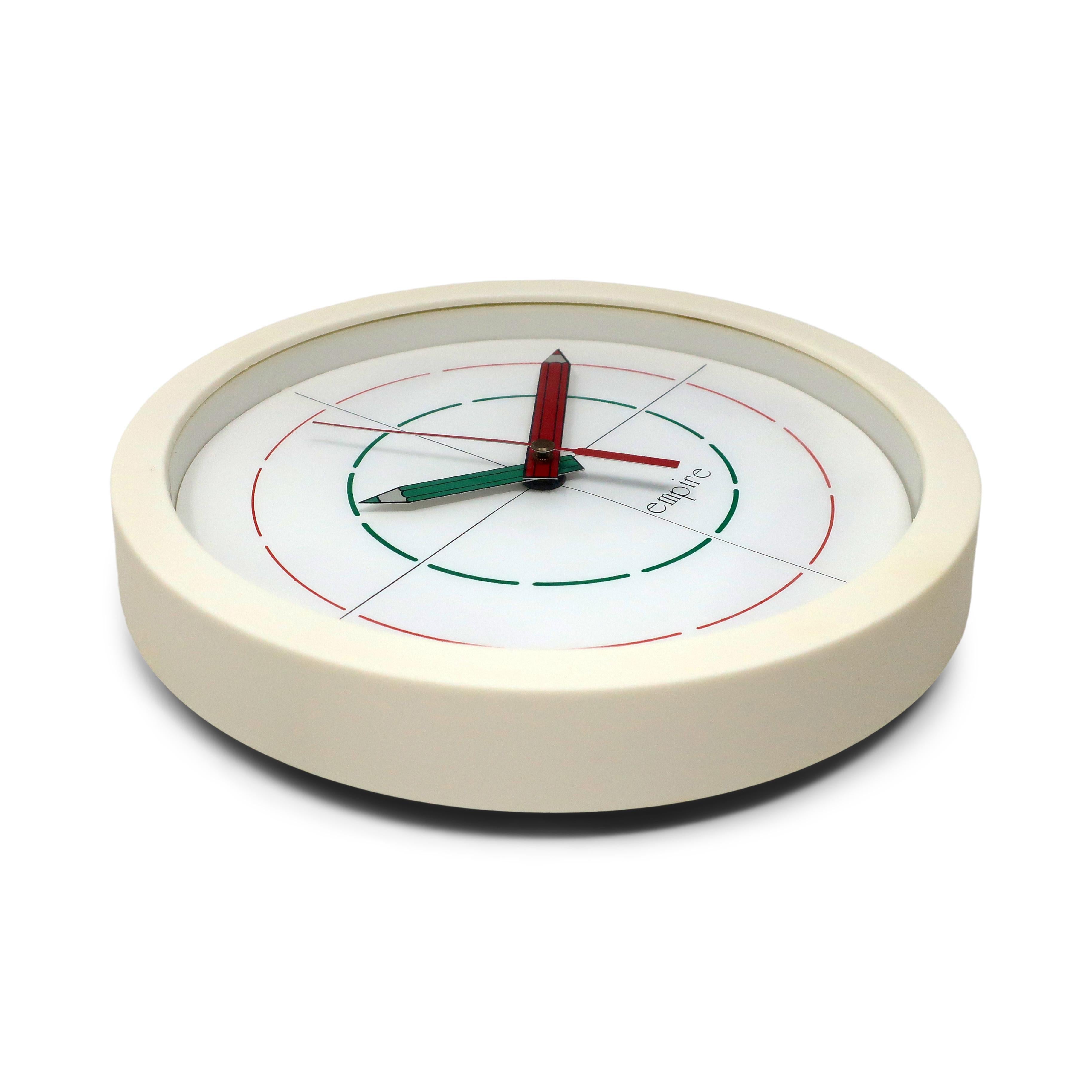 A round white 1980s wall clock by Empire. A white frame and white face with dashed red and green lines to show where the clock's numbers would appear. Hands in the shape of pencils are metal and painted red and green, with a touch of the underlying
