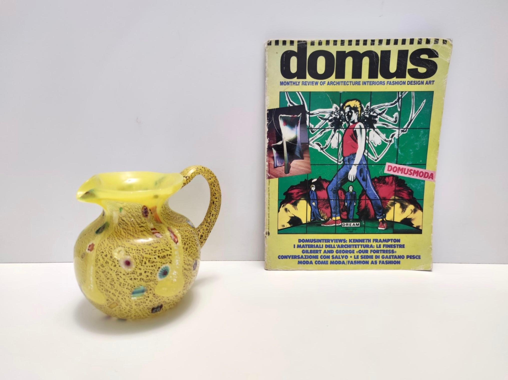 Made in Italy, anni '80.
È realizzato in vetro opalino giallo soffiato a mano con murrine e scaglie d'argento in due tonalità diverse.
Questa brocca presenta un manico a coste. 
Si tratta di un articolo vintage, quindi potrebbe presentare lievi