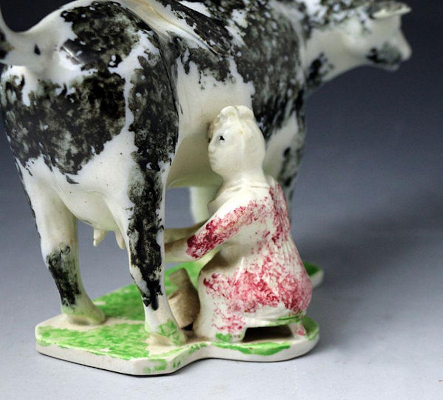 Pottery black sponge decorated cow creamer with a milking maid. The figure is modeled standing on a lime green sponge decorated base; the milkmaid is wearing a light maroon dress. The color palette and naive modeling lead to a Scottish or Northen