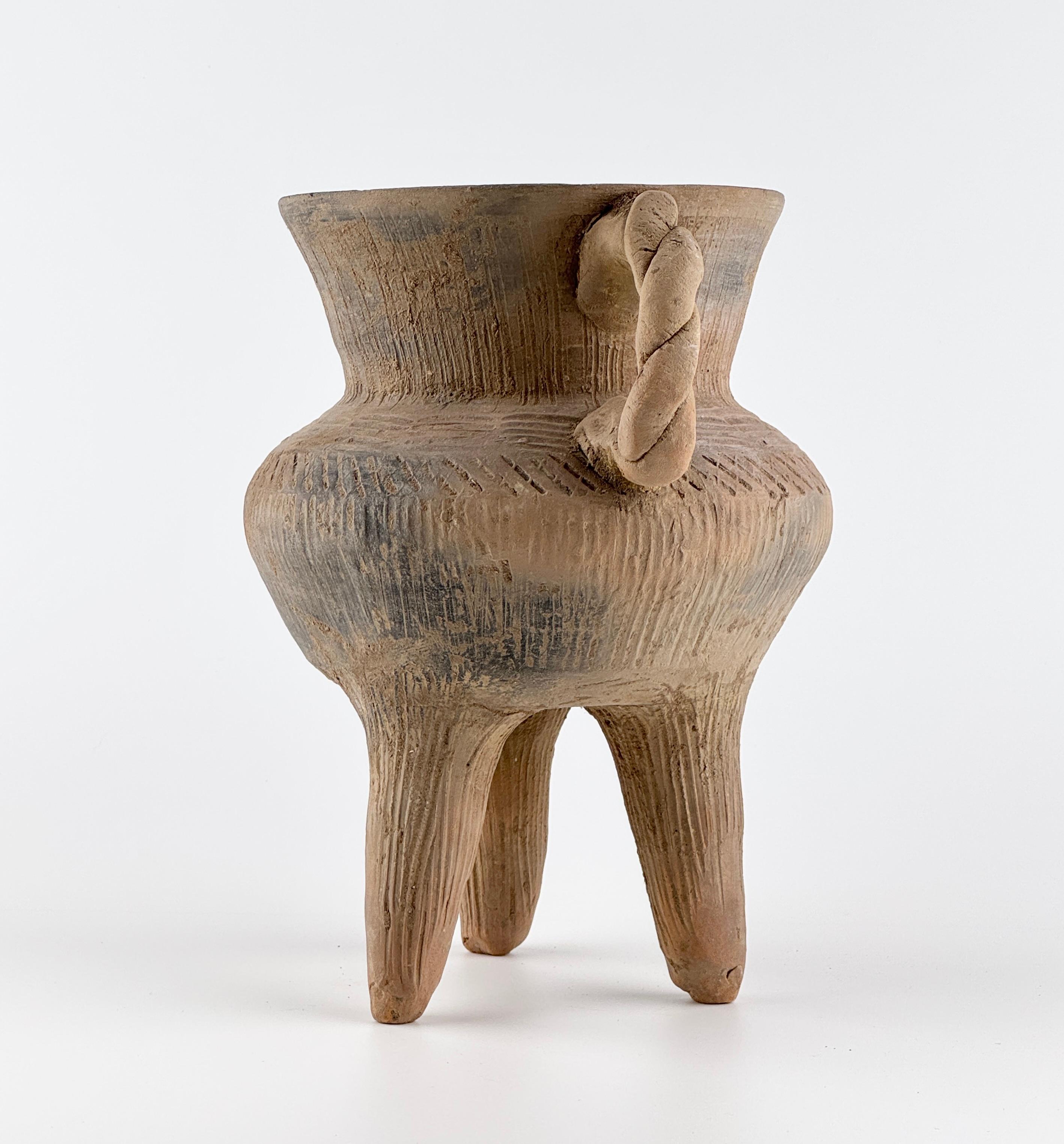The sturdy triangular legs, wide mouth, convex body, and diagonal incised patterns create a harmonious balance, showcasing a form that is both primitive and refined. Notably, the twisted handle, a rare stylistic feature of the time, adds to its