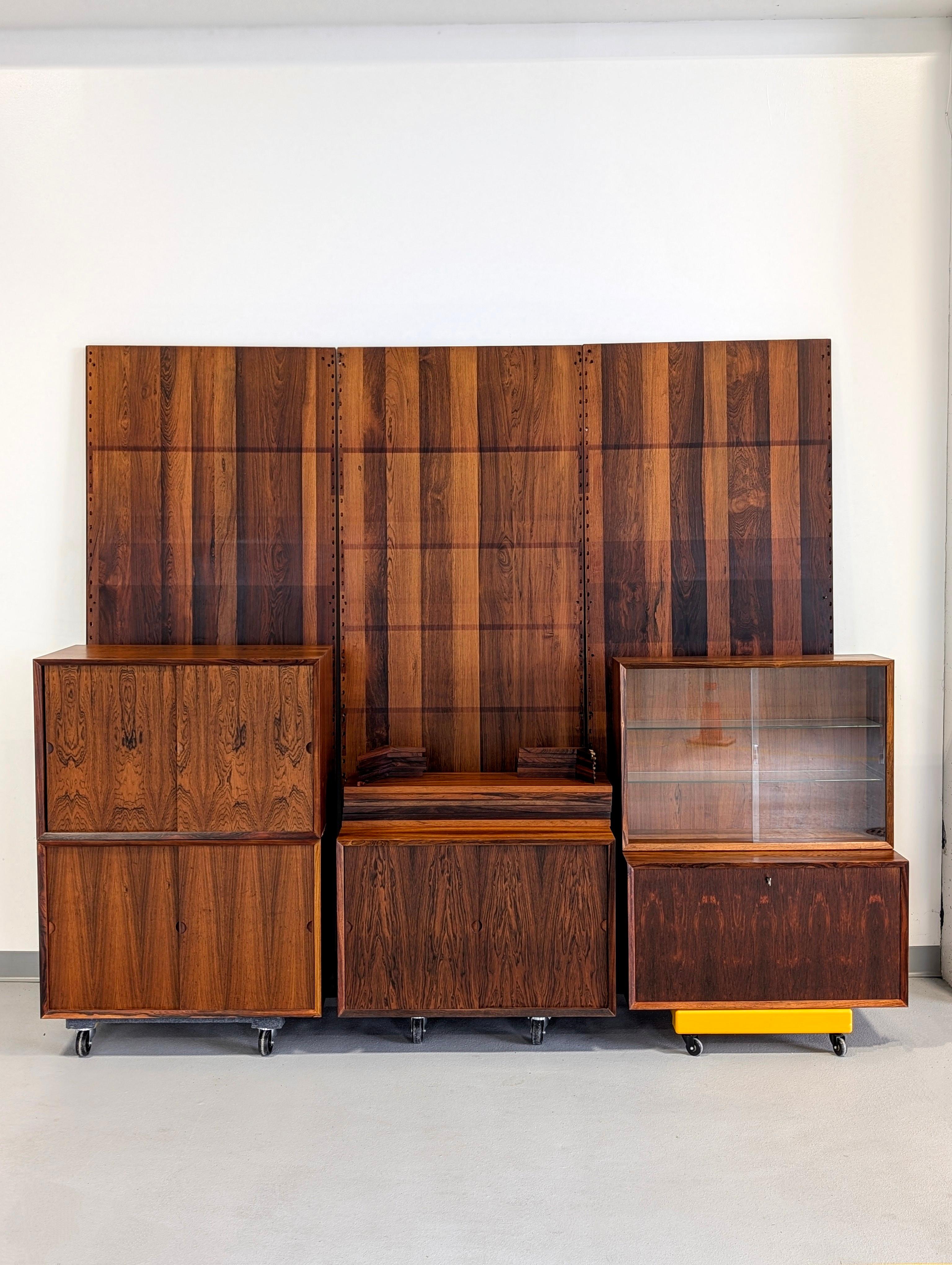  3 bay Brazilian Rosewood Royal System designed by Poul Cadovius. Features 3 wall panels, 6 shelves w/ 12 brackets, 3 large sliding door compartments, 1 glass door compartment and 1 drop down compartment w/ key. Shelves inside sliding door