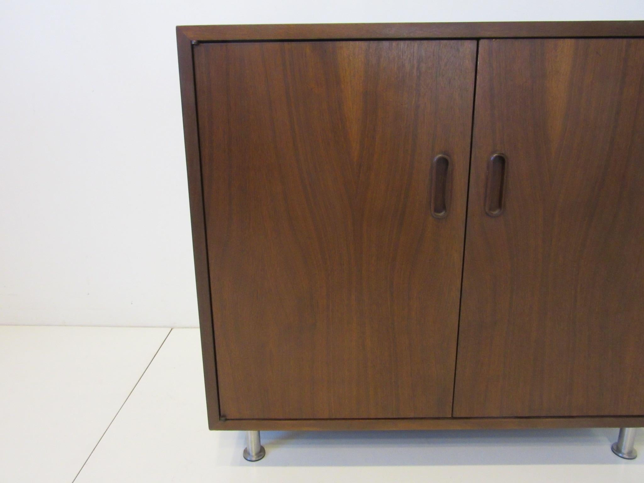 A two-door smaller dark walnut cabinet / credenza with one adjustable shelve sitting on adjustable brushed stainless steel legs, designed by Poul Cadovius for Cado made in Denmark.