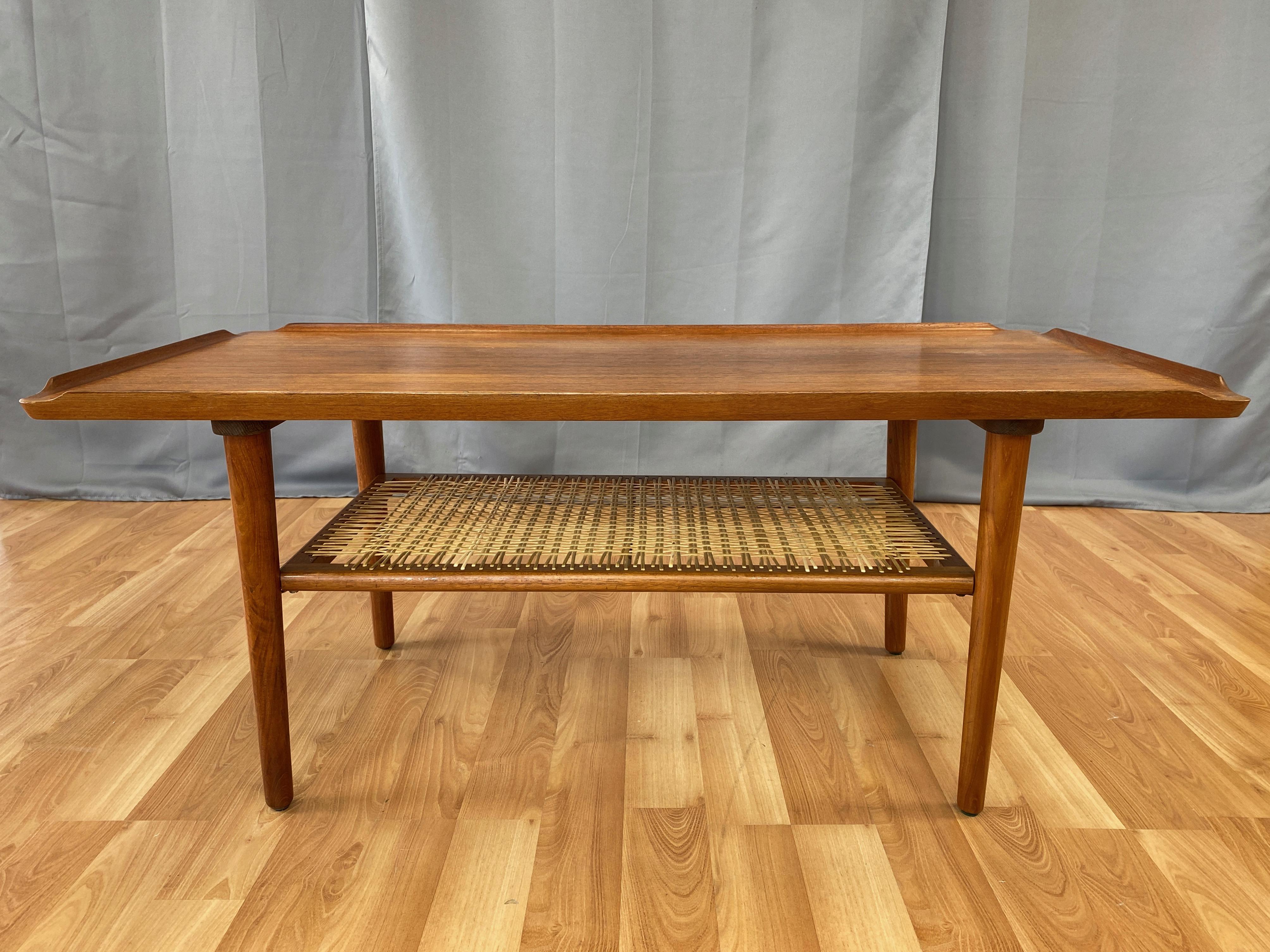 A very handsome Danish modern two-tier teak and rattan cocktail table or tall coffee table by Poul Jensen for Selig.

Top of bookmatched teak with sculptural raised lips on all four sides. Below is a shelf of open-weave rattan in a teak frame.
