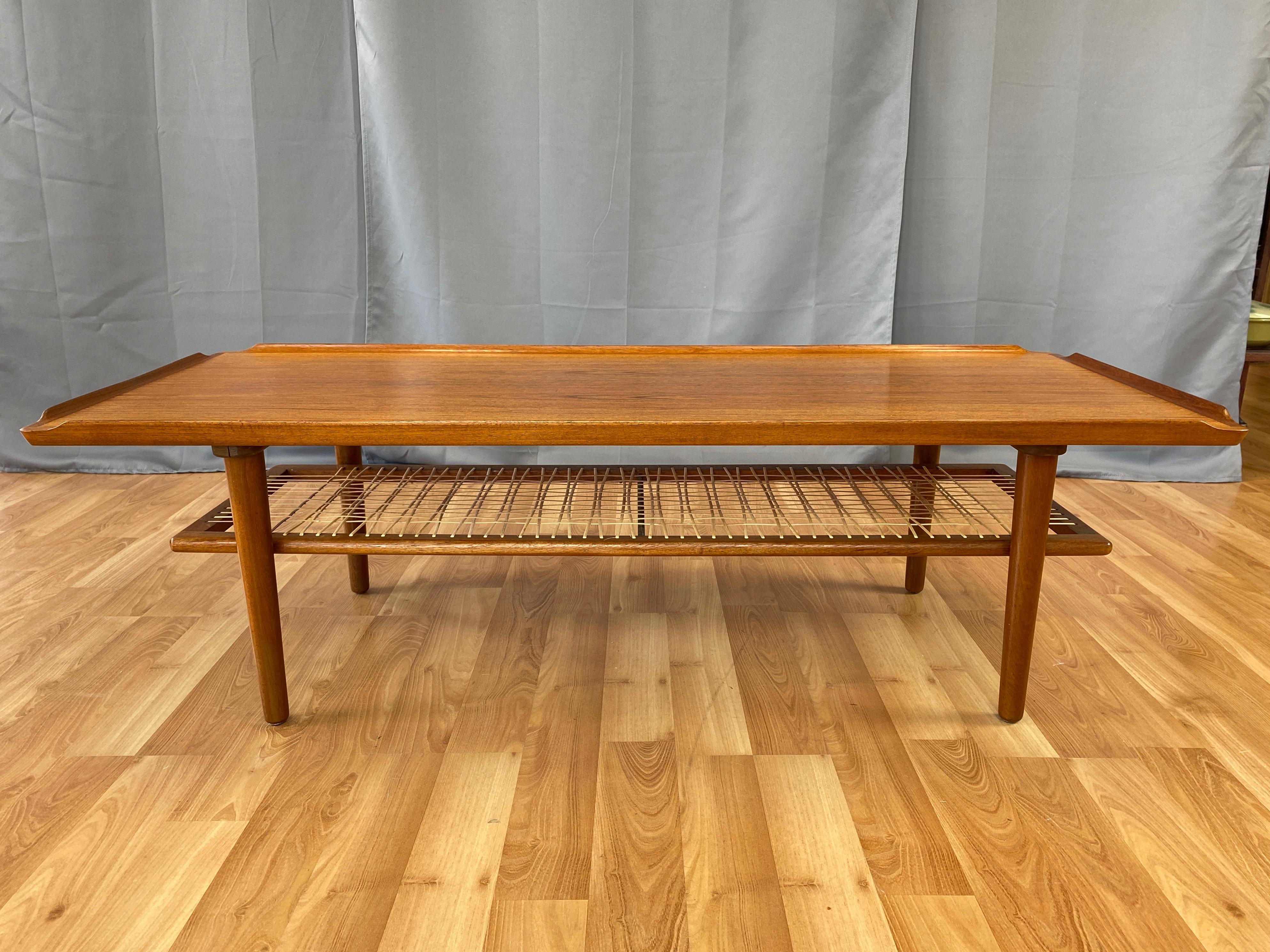 A very handsome Danish modern two-tier teak & rattan coffee table by Poul Jensen for Selig.

Expansive top of bookmatched teak with sculptural raised lips on all four sides. Below is a shelf of open-weave rattan in a teak frame. Stands on slightly