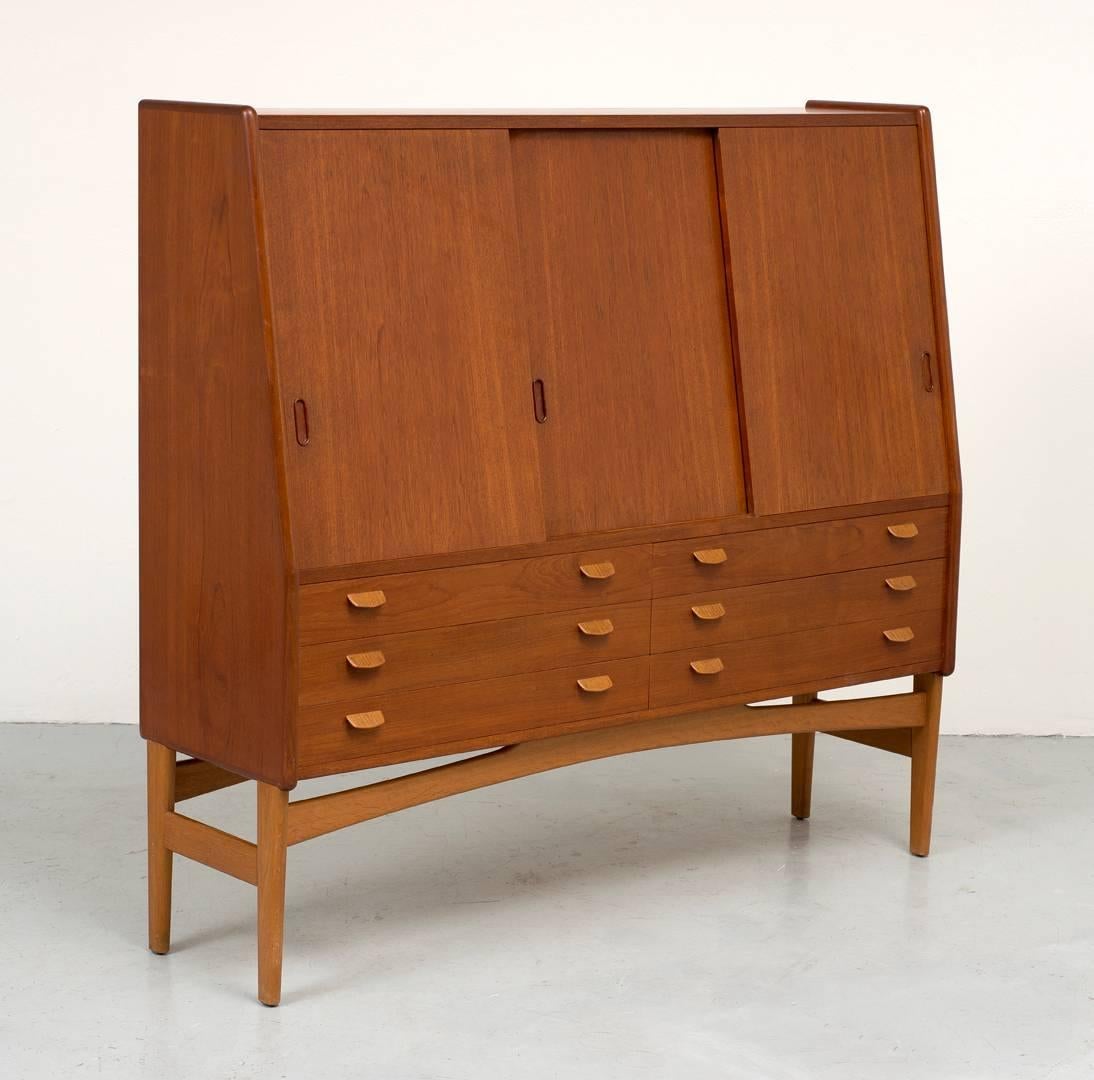 A tall teak cabinet with six drawers below with tab pulls, and three sliding panels on top revealing oak shelves.
