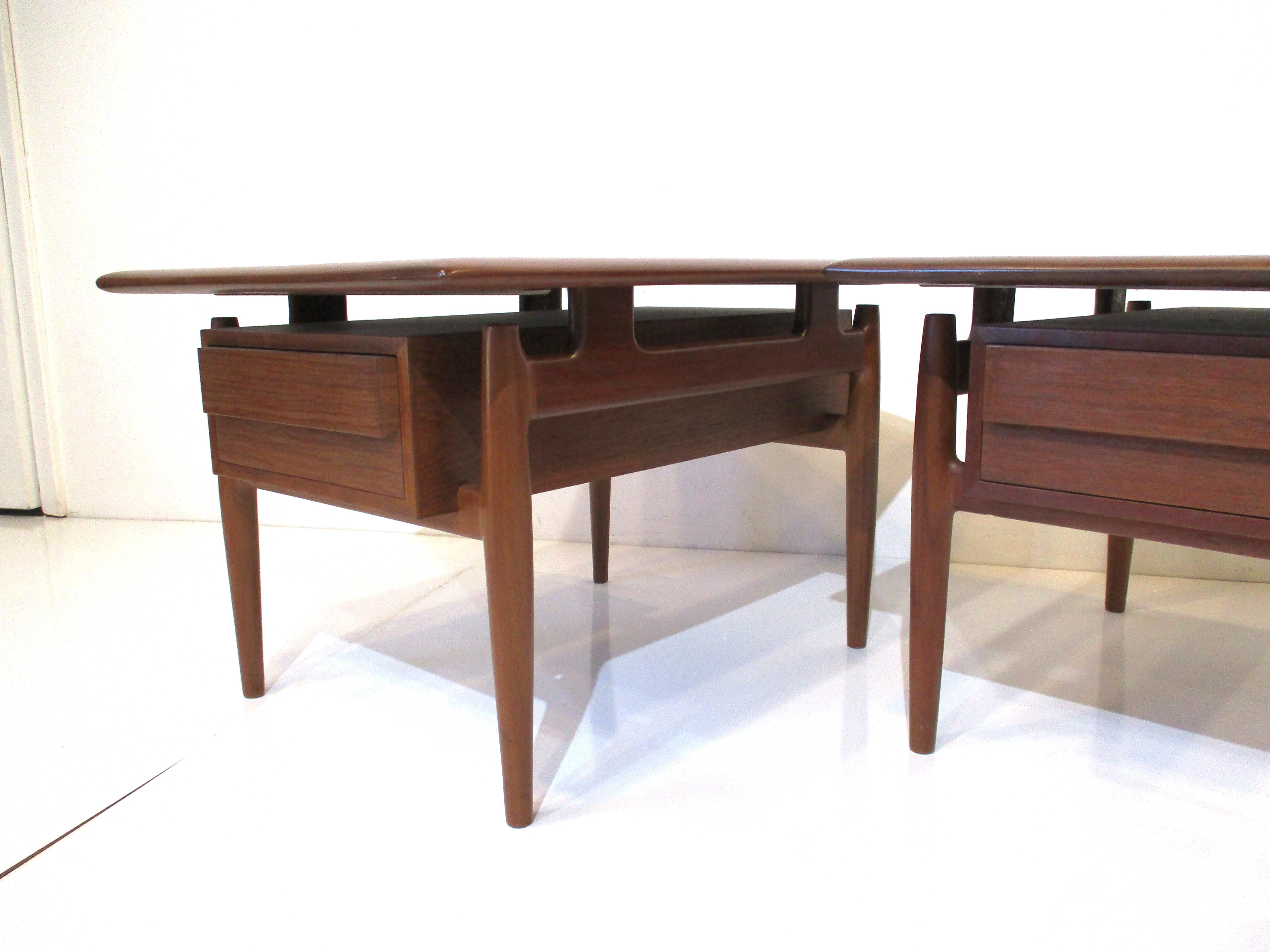 A pair of very well crafted teak side tables with floating top, single drawer to each with protruding beveled handle across the front of the drawers and open backside opposite the drawer for storage. The sculptural side mounted leg structure is