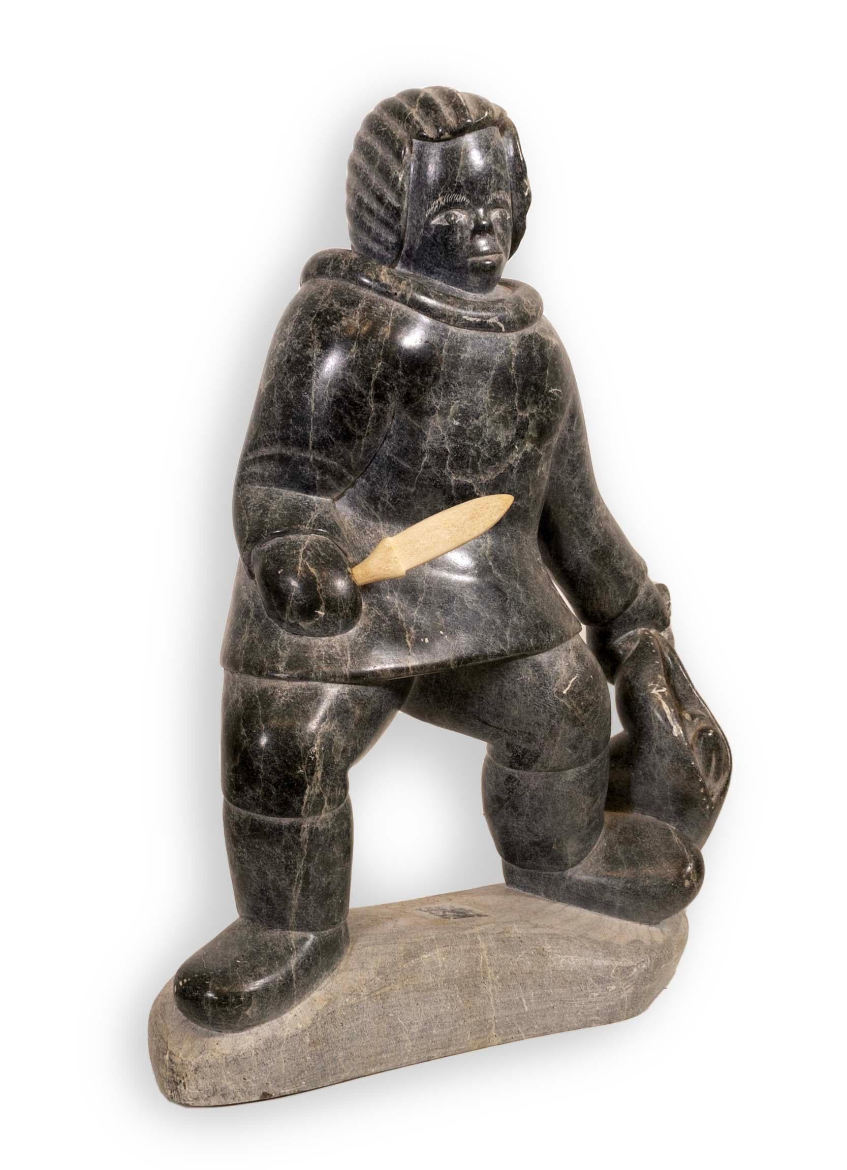An Inuit figurative soapstone carving sculpture by the Povungnituk Cooperative. From the Quebec region in the Canadian Artic. Circa 1980s. The composition is of a male hunter holding a knife and small animal. ThePovungnituk Cooperative is a group of