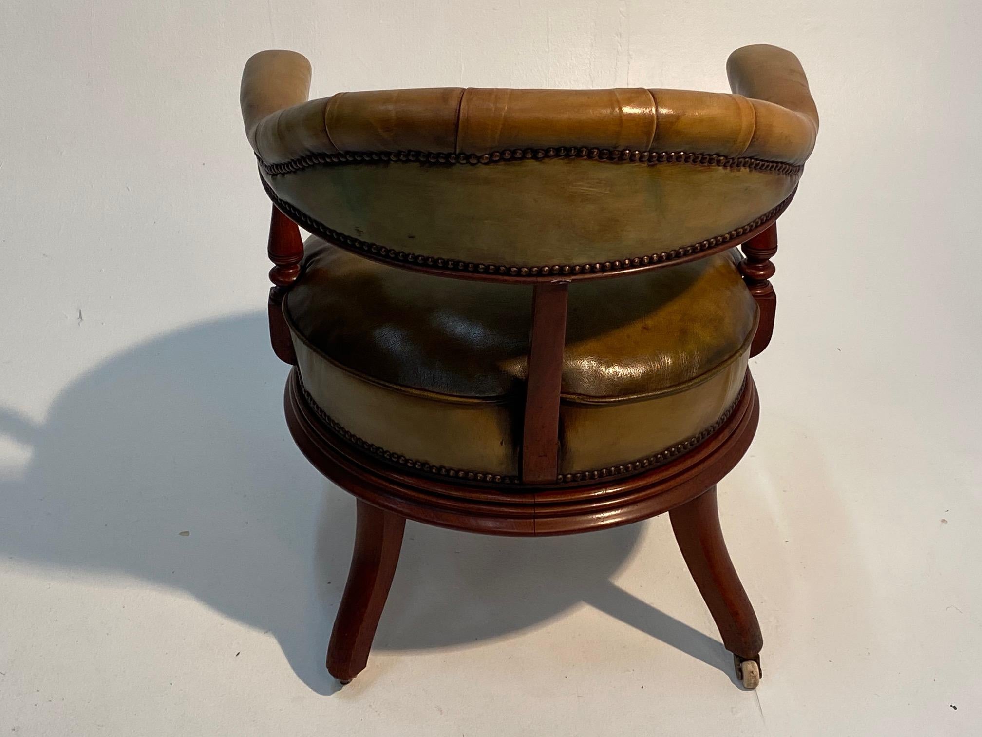 A very handsome rotating library or desk chair having mahogany frame with curved back and button tufted backrest, stylish pill box round hat shaped seat cushion, upholstered in mossy green leather with brass nailheads. Legs have original porcelain
