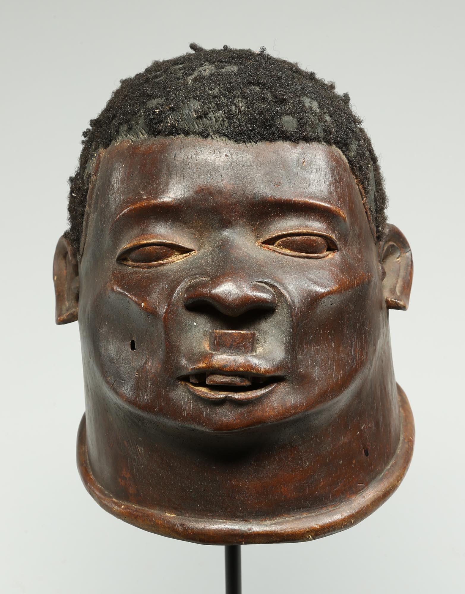 Powerful portrait of a Makonde man with a traditional lip plug in his upper lip, high carved cheek bones in the form of a finely carved helmet mask, with attached hair on top from Tanzania, East Africa. Early 20th century with dark brown patina from