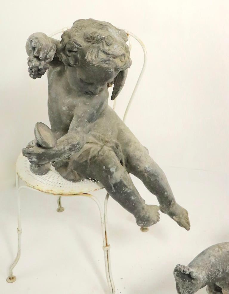 Stunning pair of cast lead cupids, originally part of a grand public fountain, now destroyed. The seated cupids hold a wine goblet in one hand, and a cluster of grapes on the other. One figure has some damage, specifically the wine glass is bent,