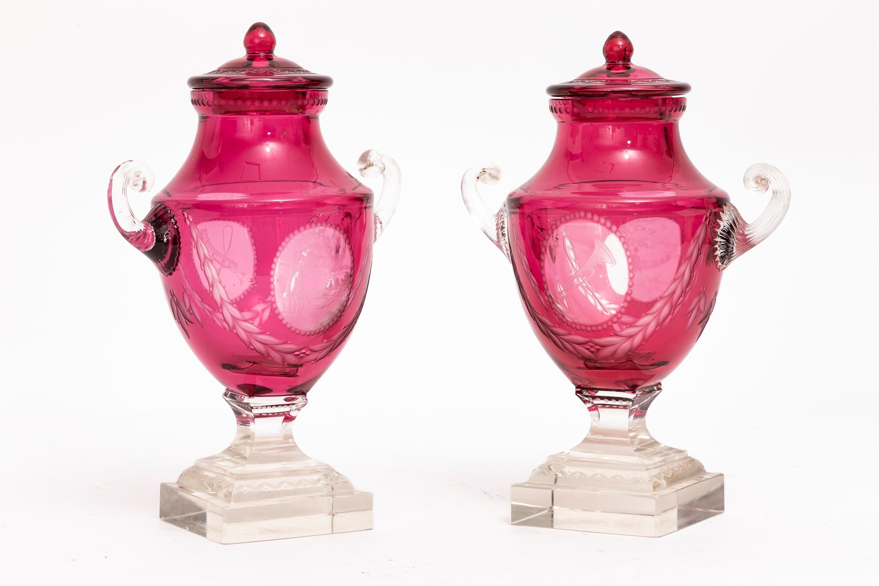 A Magnificent Pair of Covered Crystal Double Overlay Cranberry Cut-to-Clear Hunting Trophy Vases with applied shell handles and stepped bases, Attributed to Baccarat. These vases are characterized by their rich cranberry hue, meticulously etched and