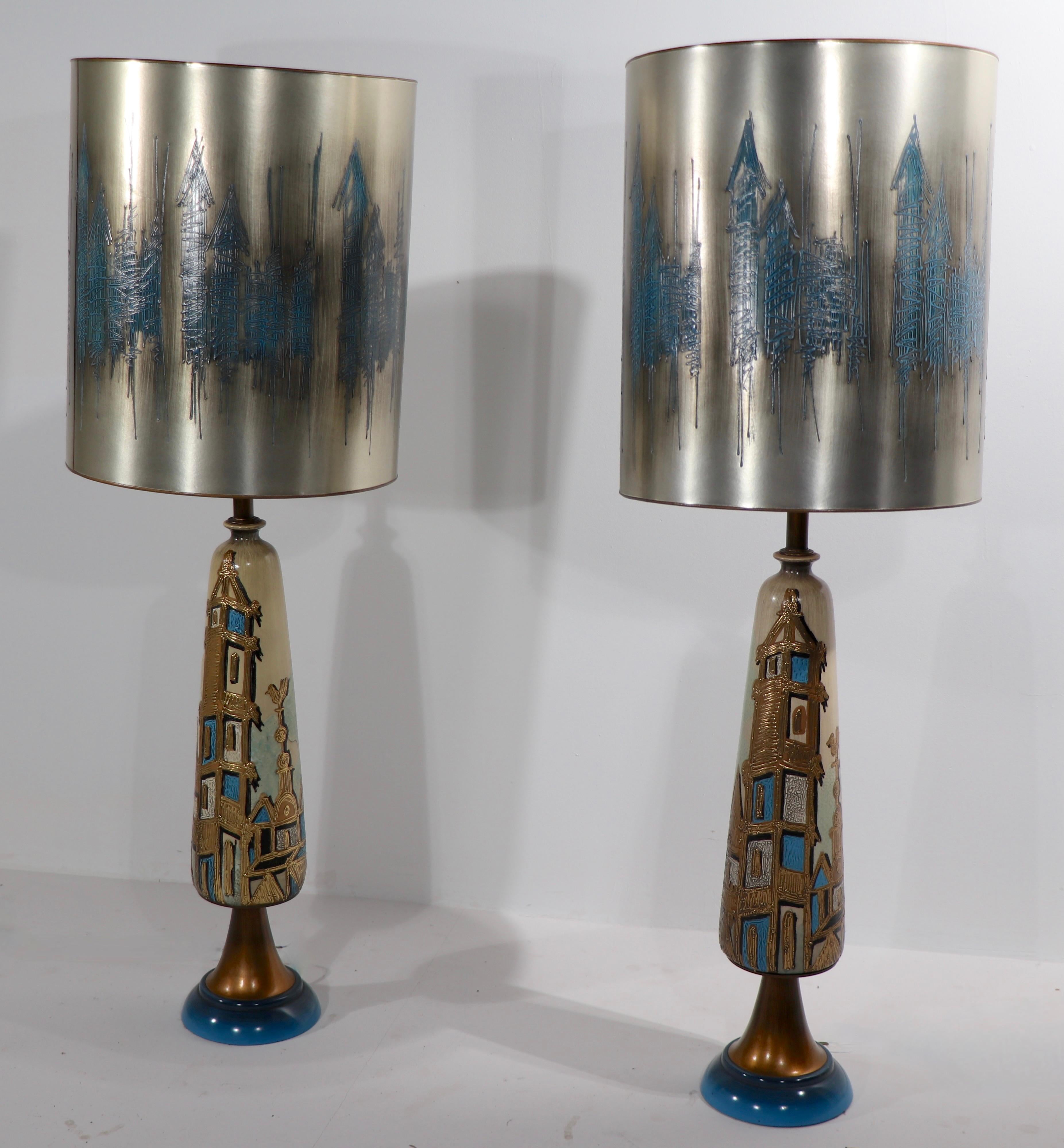 Incredible pair of Brutalist, mid century, Hollywood Regency, table lamps, with original silver and blue decorated shades. The lamps depict a stylized city scene with columns, towers and buildings. The ceramic bodies have an off white and tan tone